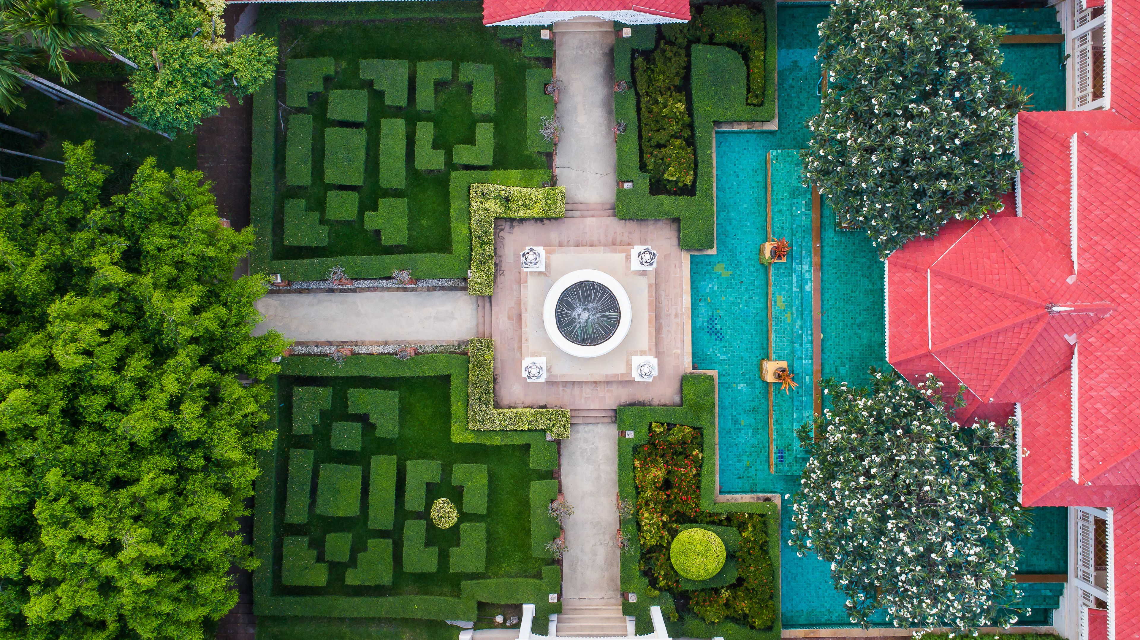 Eine Luftaufnahme eines Innenhofs mit einer zentralen Fontäne, umgeben von geometrisch angelegten Blumenbeeten und einem Pool.