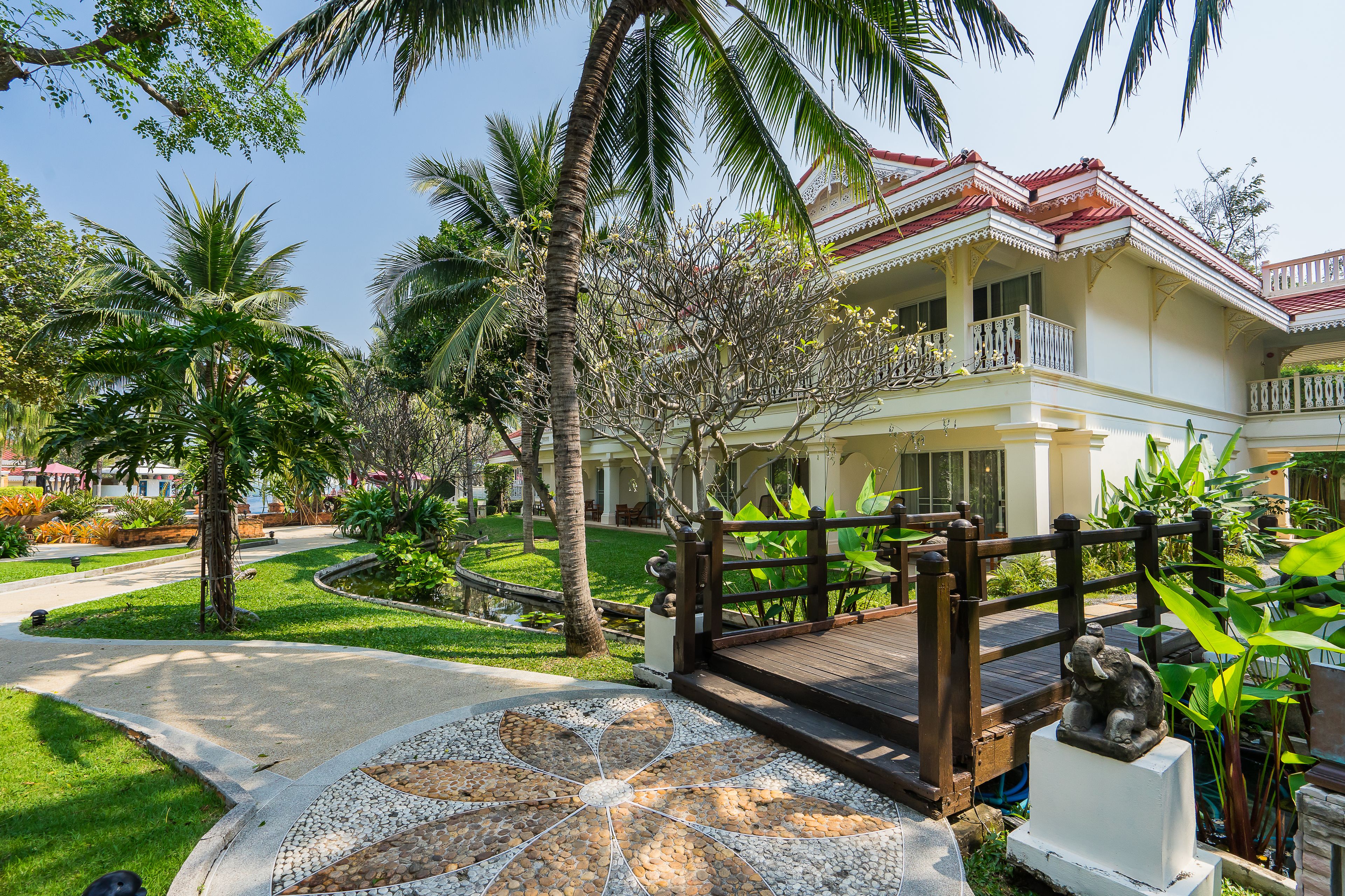Un resort tropical con un camino pavimentado que conduce a un puente de madera, rodeado de exuberante vegetación y palmeras.