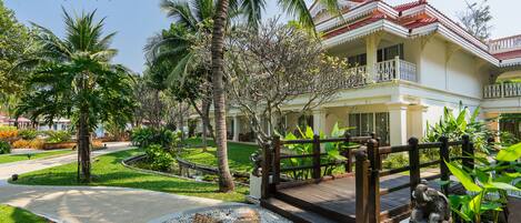 Un resort tropical con un camino pavimentado que conduce a un puente de madera, rodeado de exuberante vegetación y palmeras.