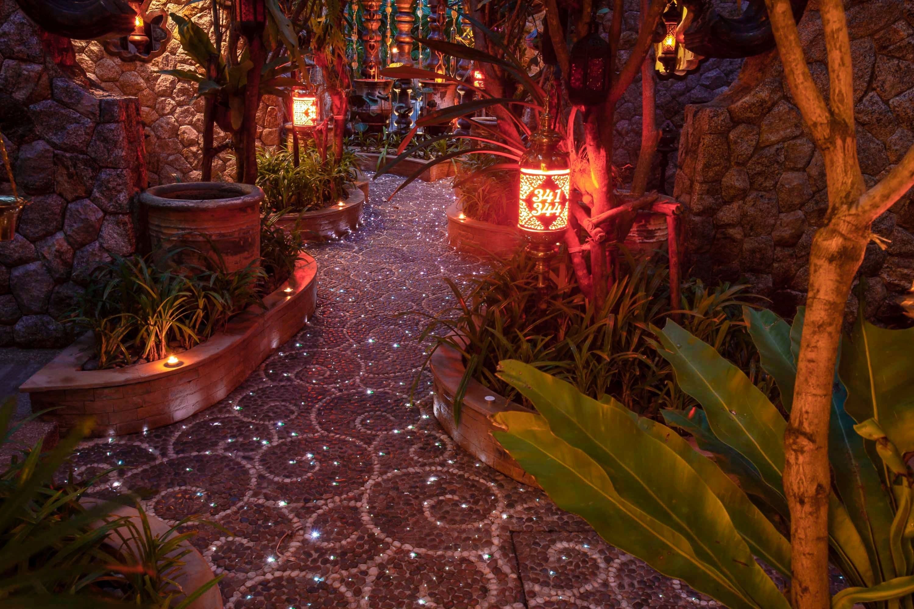 A stone pathway leading through a tropical garden with potted plants and decorative lanterns.