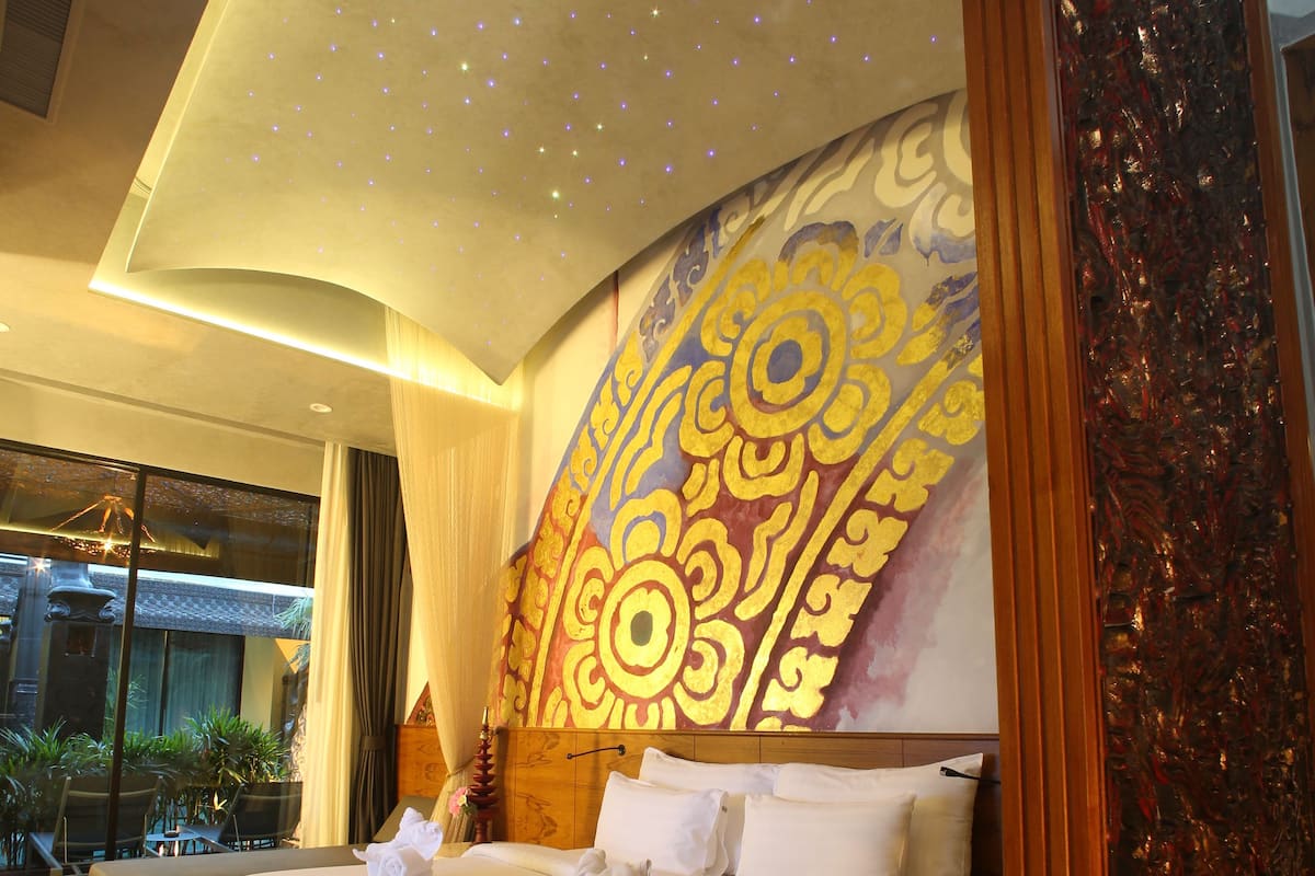 Una cama grande con cabecera de diseño dorado, suelo de madera y vistas a un edificio a través de amplios ventanales.