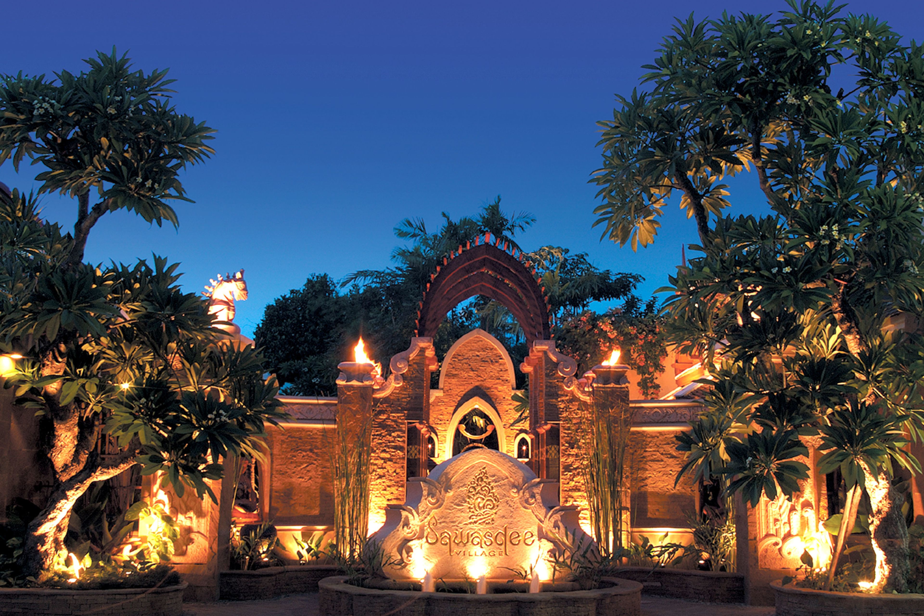 Una entrada luminosa con un arco de piedra, palmeras y una fuente decorativa.