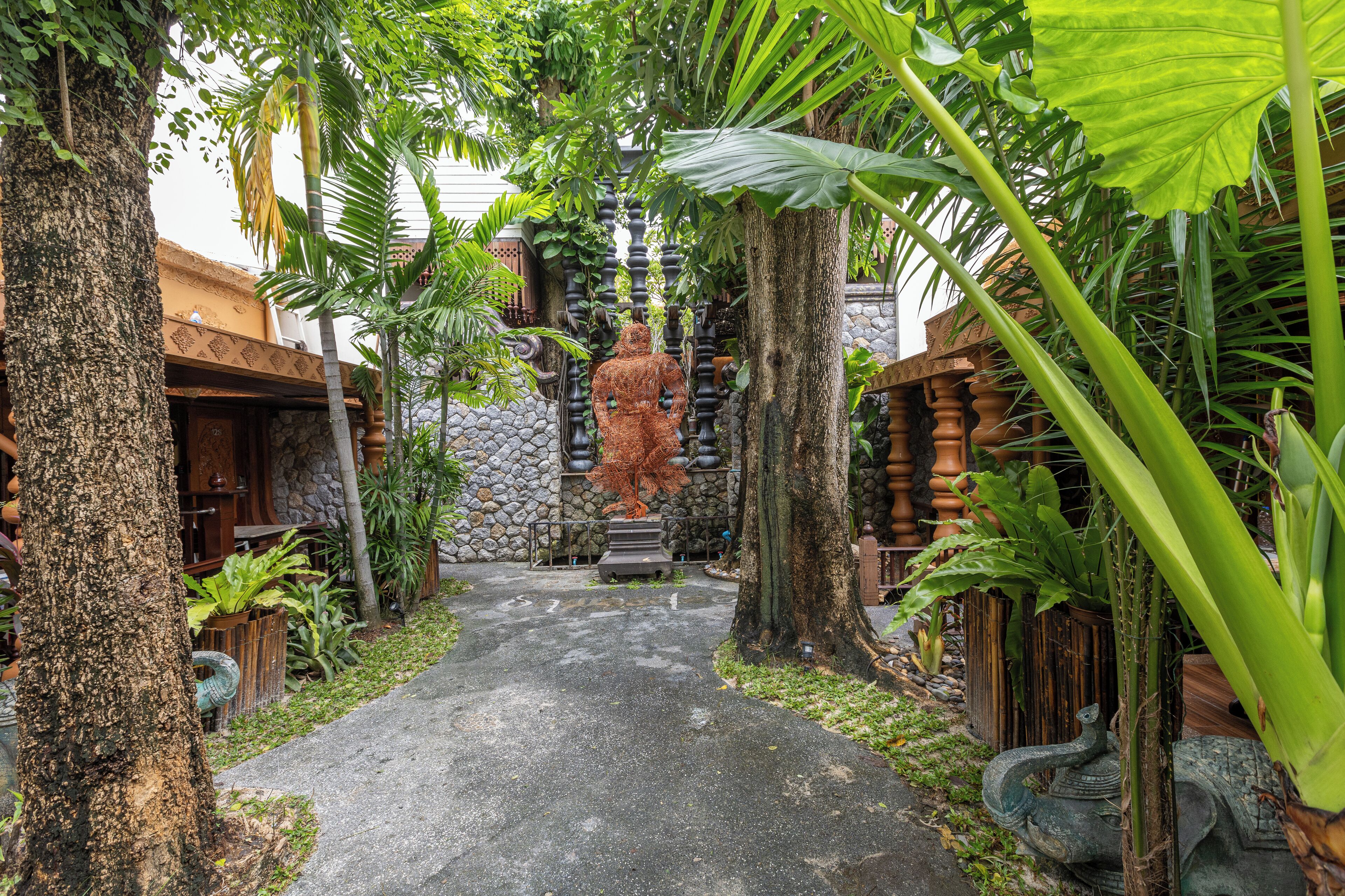 A narrow pathway flanked by lush tropical plants and a stone wall with a statue.