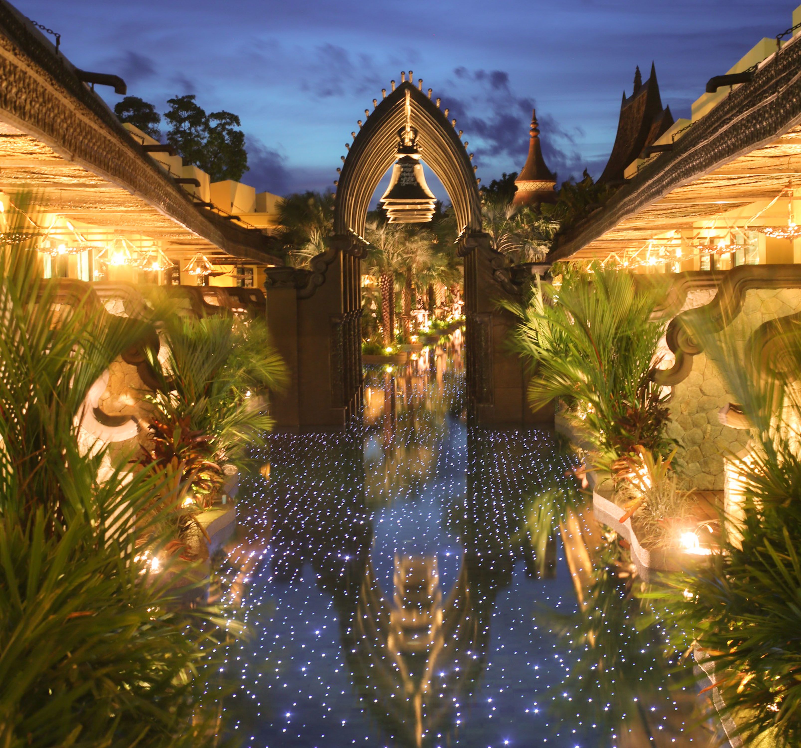 A nighttime view of a courtyard with a reflective water surface, ornate architecture, and palm trees.