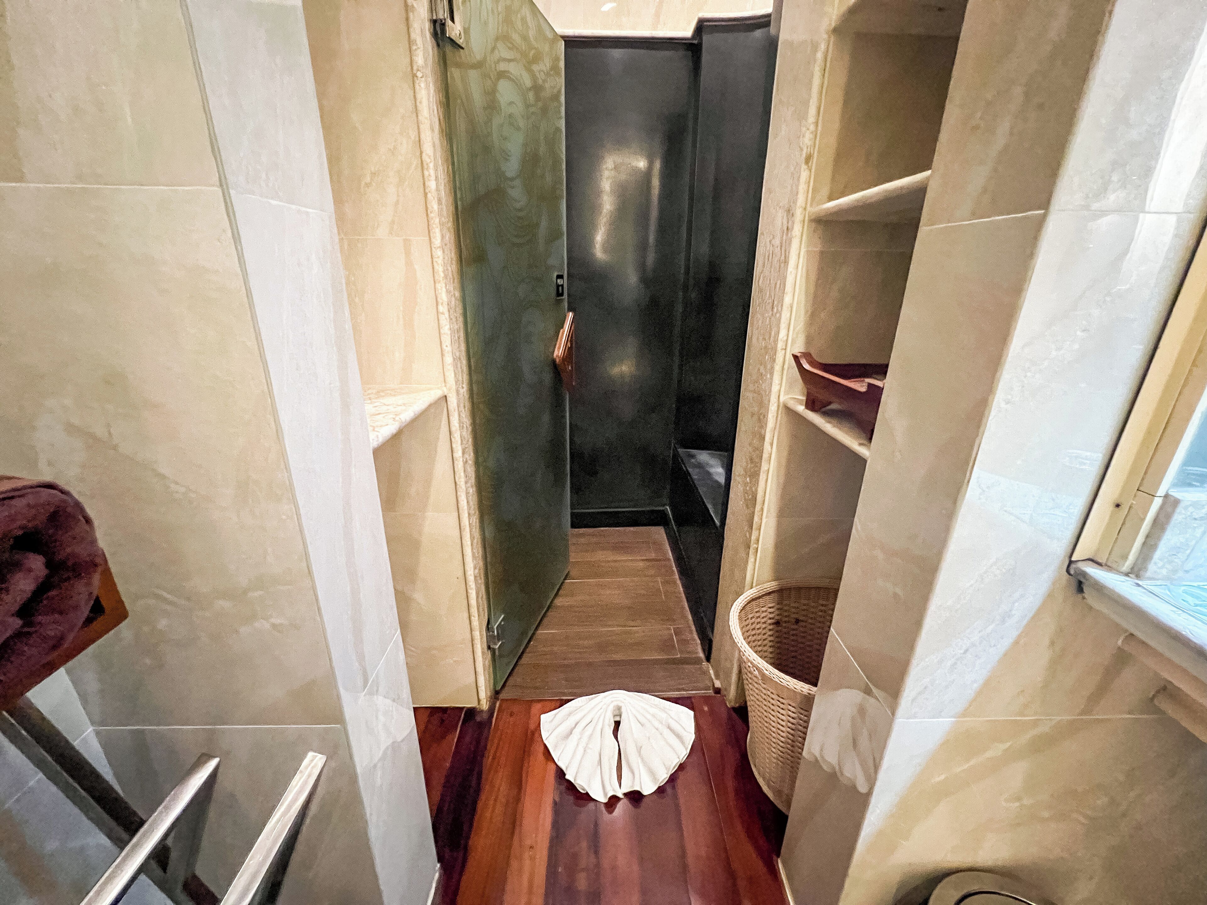 A narrow bathroom with a wooden floor, a white towel on the floor, a wicker basket, and a towel rack.