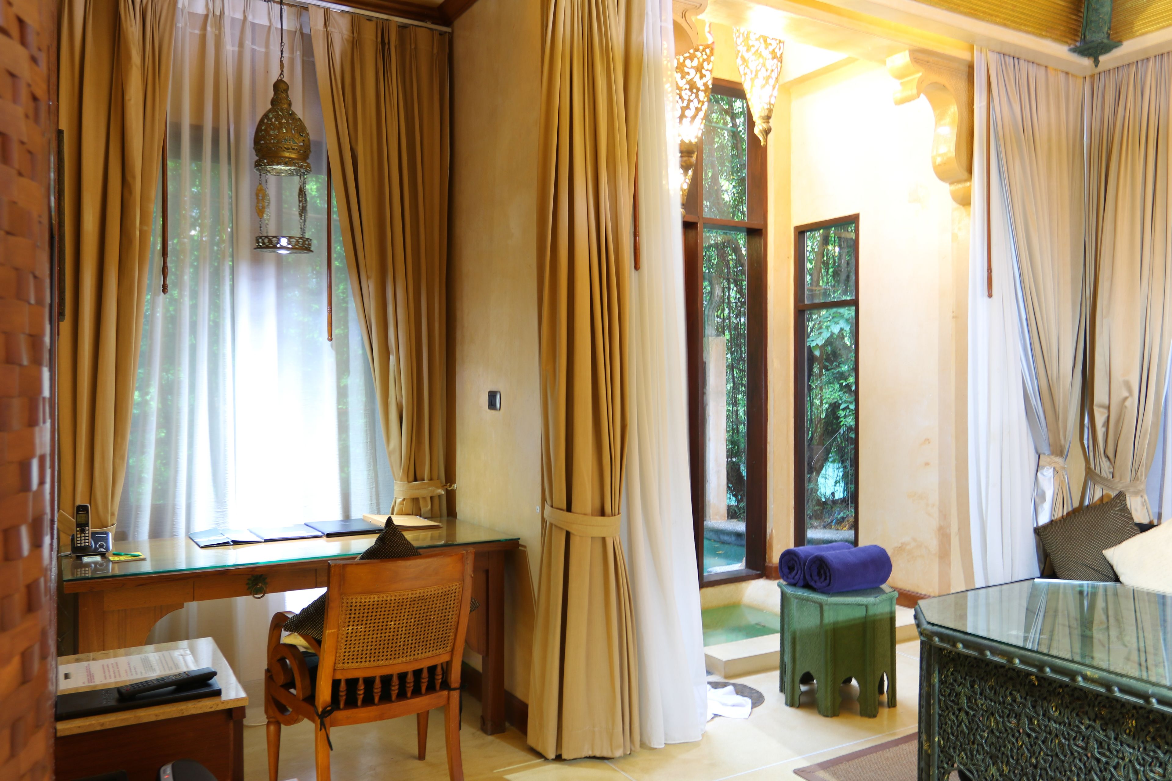 Una habitación con un escritorio de madera, una silla, una mesa de vidrio, una lámpara colgante y vistas a zonas verdes al exterior.