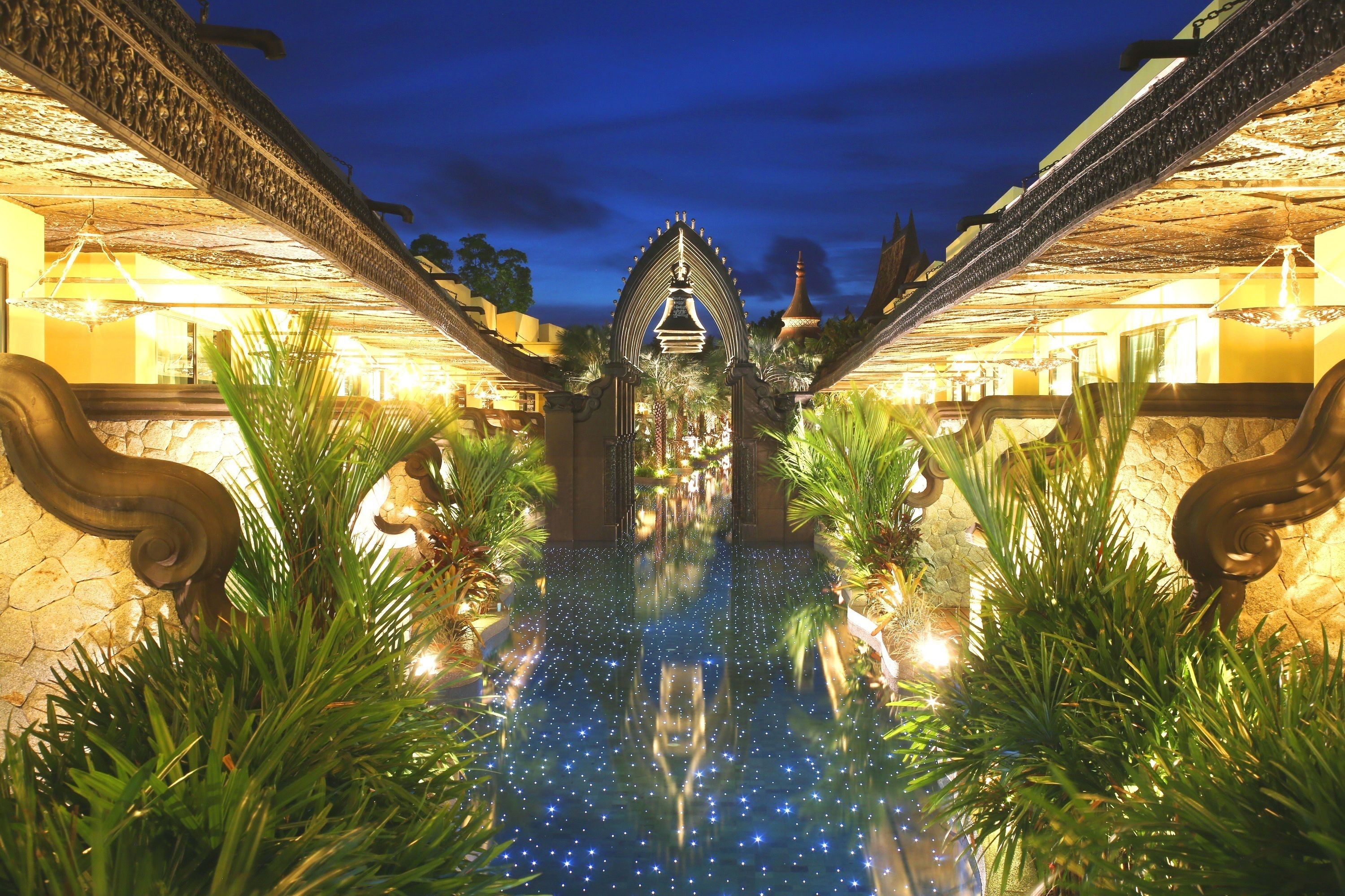 Vista nocturna de la entrada de un hotel con un arco decorativo, una piscina reflectante y senderos bien iluminados.