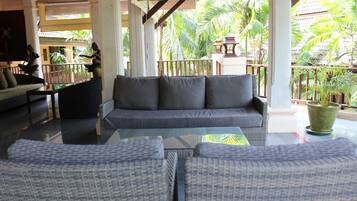 A spacious lounge area with a grey sofa and wicker chairs, a glass coffee table, and a view of tropical plants and a traditional building in the background.