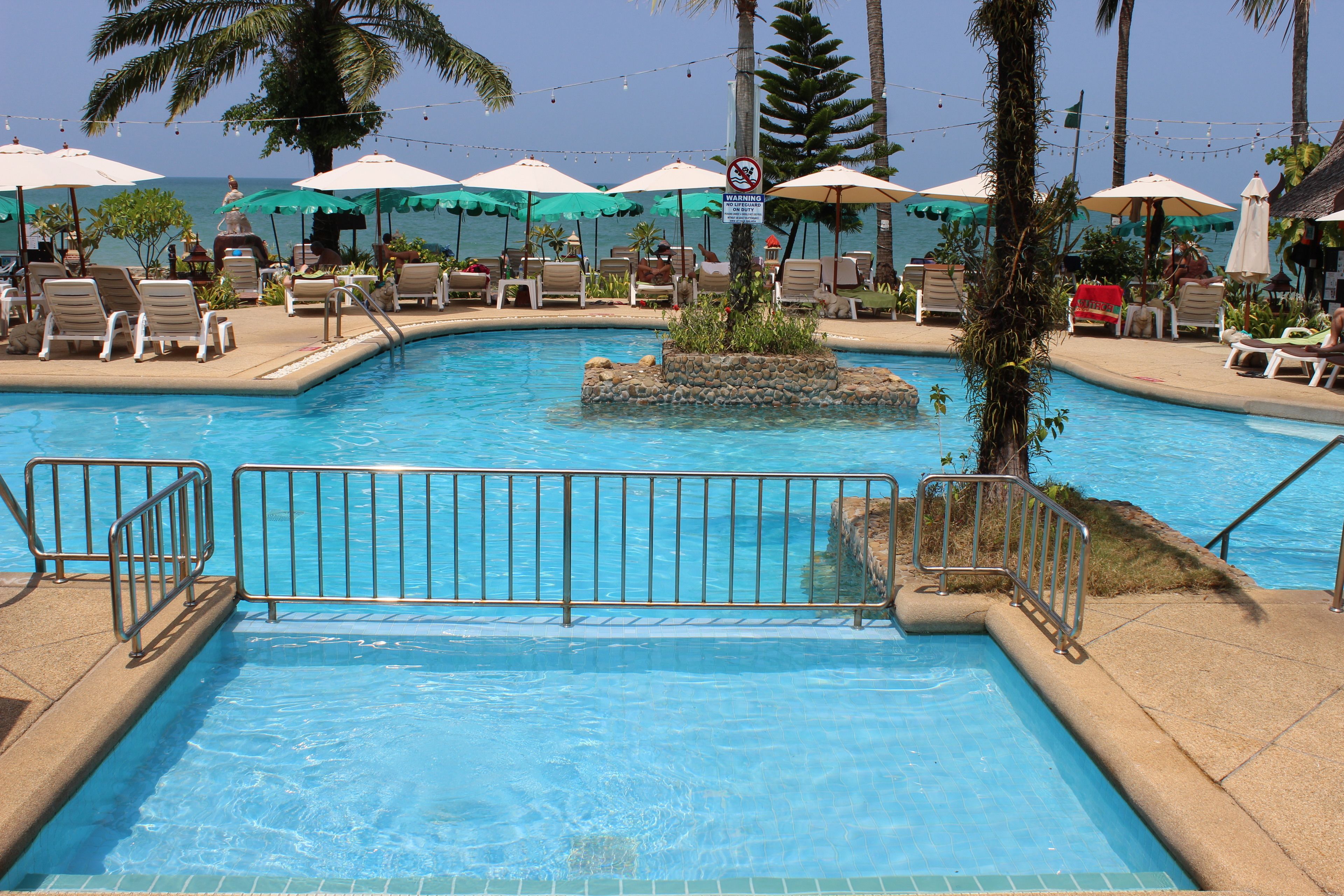 A swimming pool with lounge chairs and umbrellas, surrounded by palm trees and a view of the ocean.