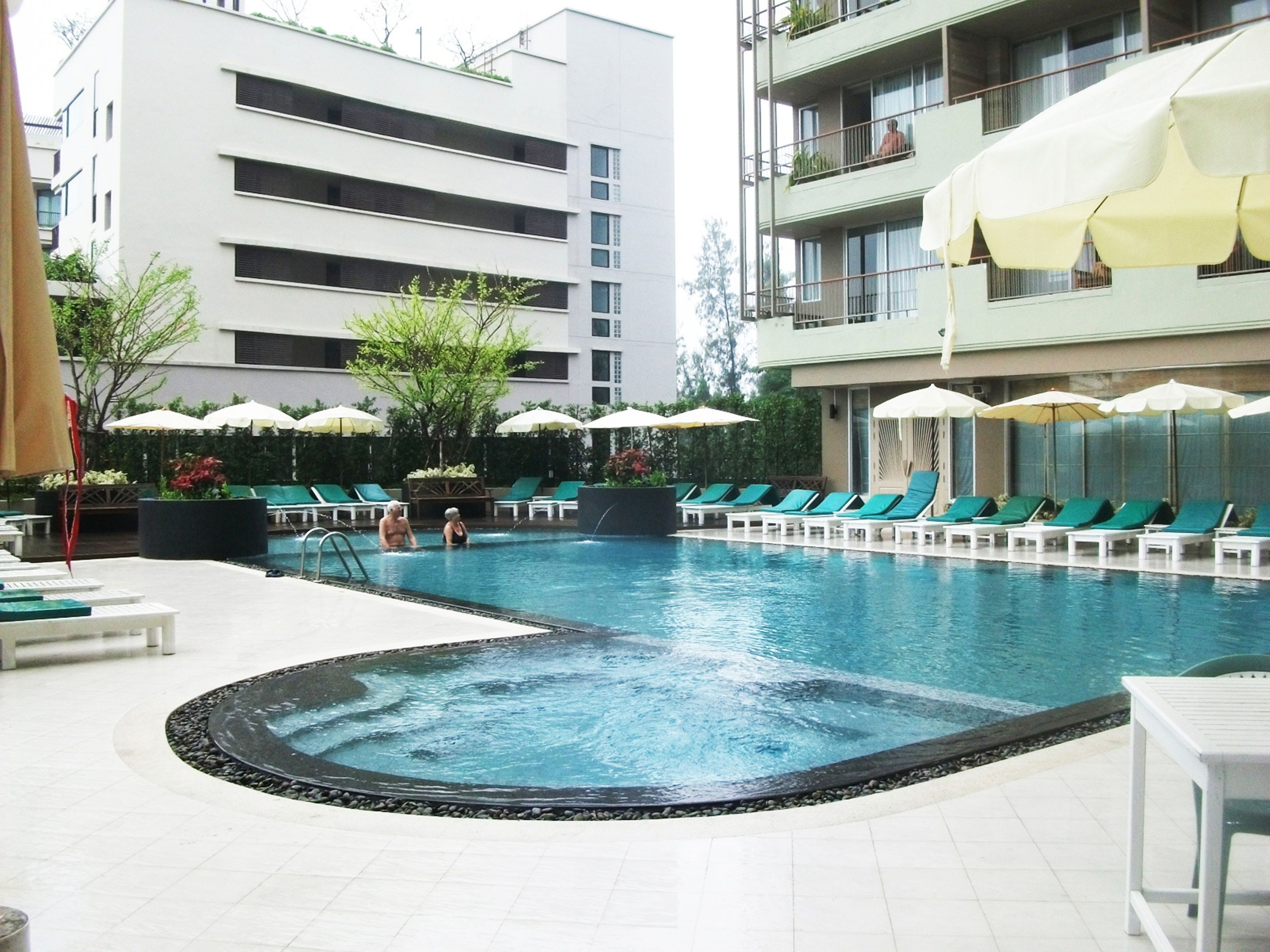 A swimming pool with lounge chairs and umbrellas, surrounded by buildings.