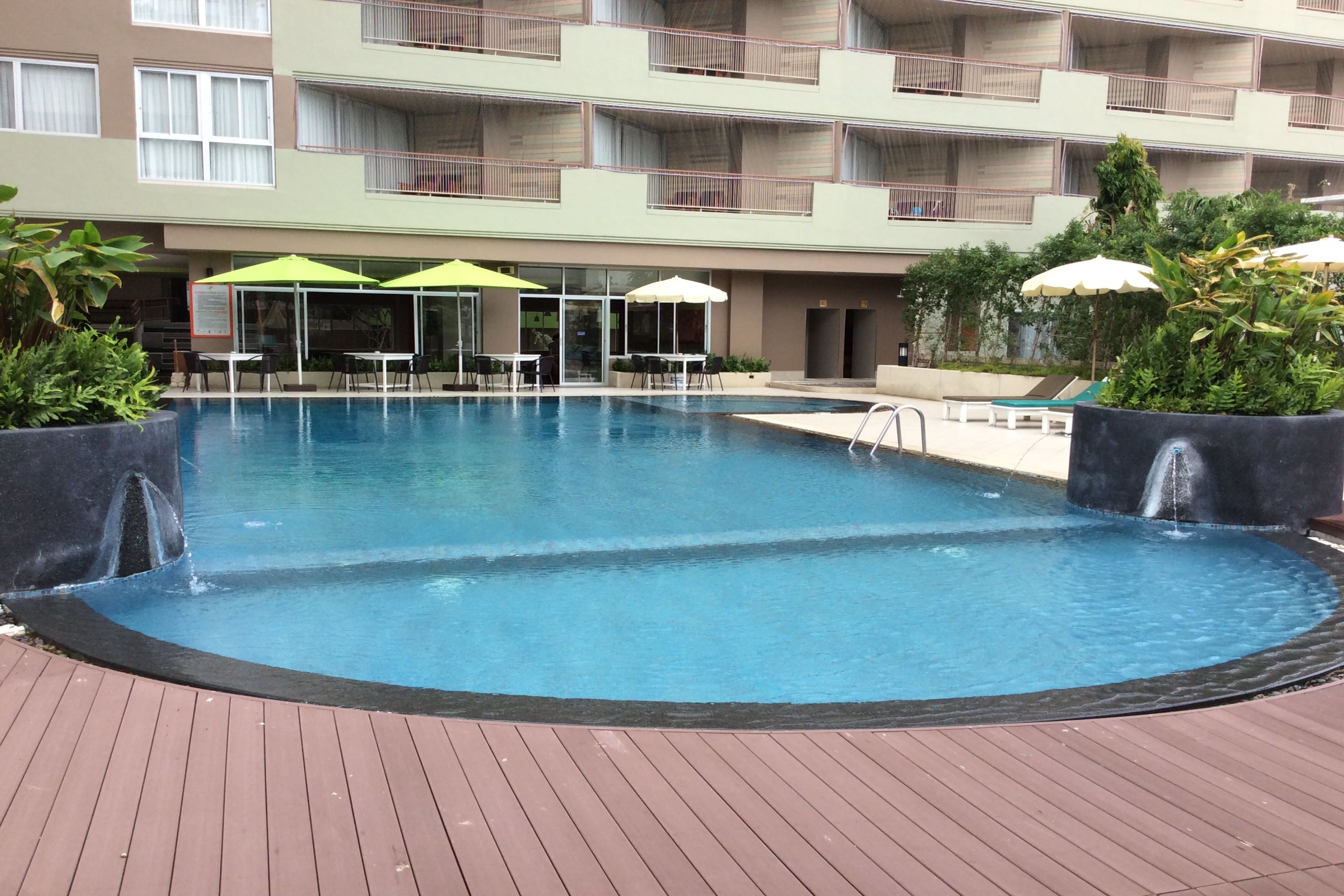 A swimming pool with a wooden deck, surrounded by lounge chairs and umbrellas, and a multi-story building in the background.