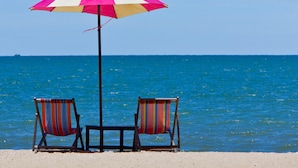 A beach scene with a pink and white umbrella, two striped beach chairs, and a clear blue sky. - Long Beach Cha-Am Hotel (Cha-am)