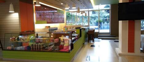 A brightly lit indoor space with a green counter displaying snacks and beverages, a pool bar area, and large windows offering a view of an outdoor swimming area.