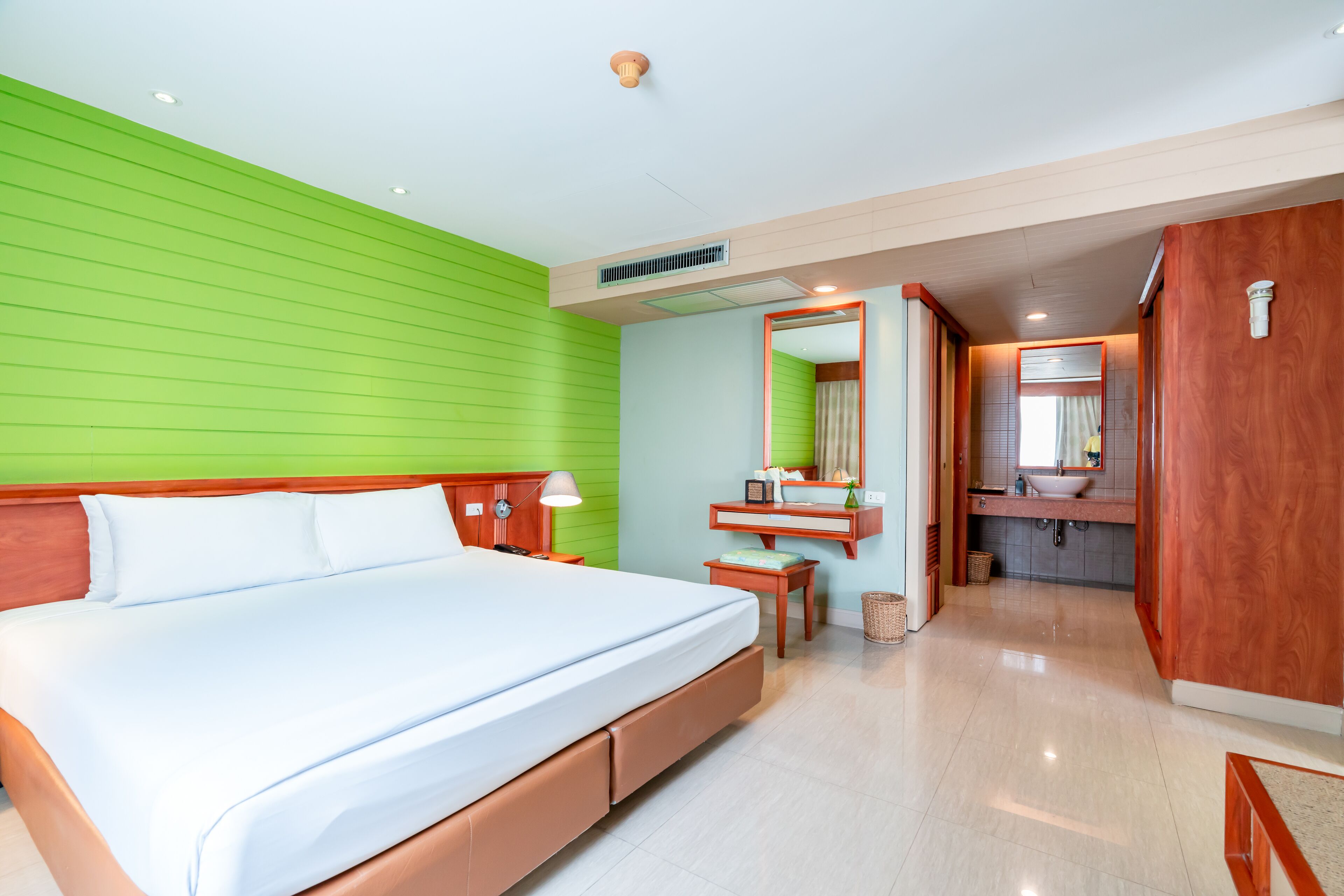 A modern hotel room with a large bed, a wooden headboard, a green accent wall, a vanity with a mirror, and a bathroom with a sink and toilet.