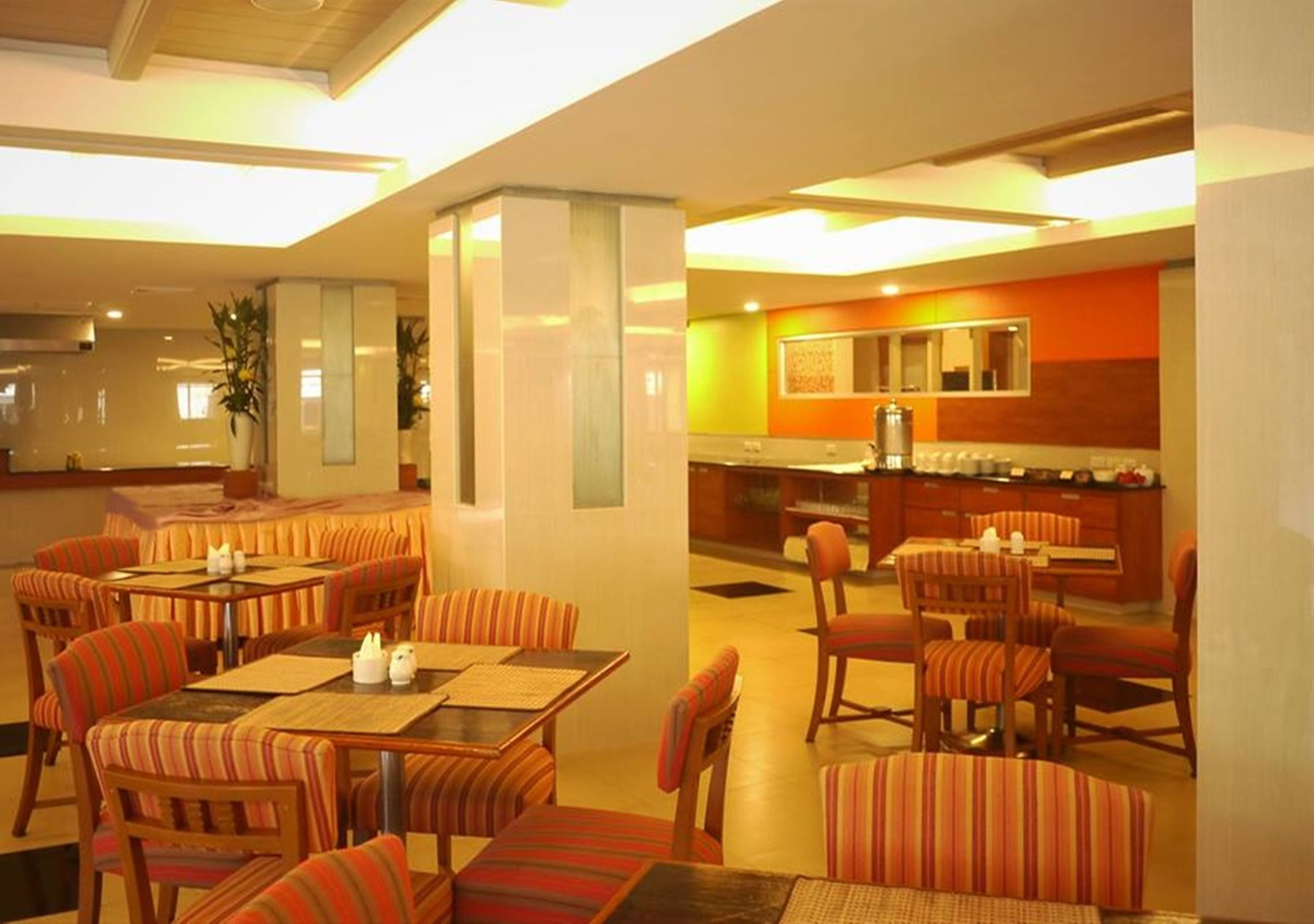 A hotel dining area with striped upholstered chairs and tables, a central pillar, and a kitchen area with a counter and stools.