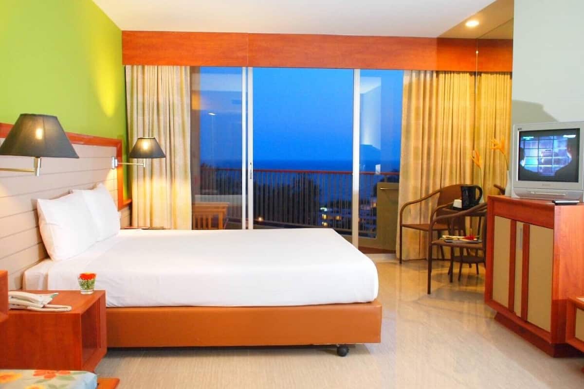 A hotel room with a bed, a desk, a chair, a TV, and a balcony with a view of the city.