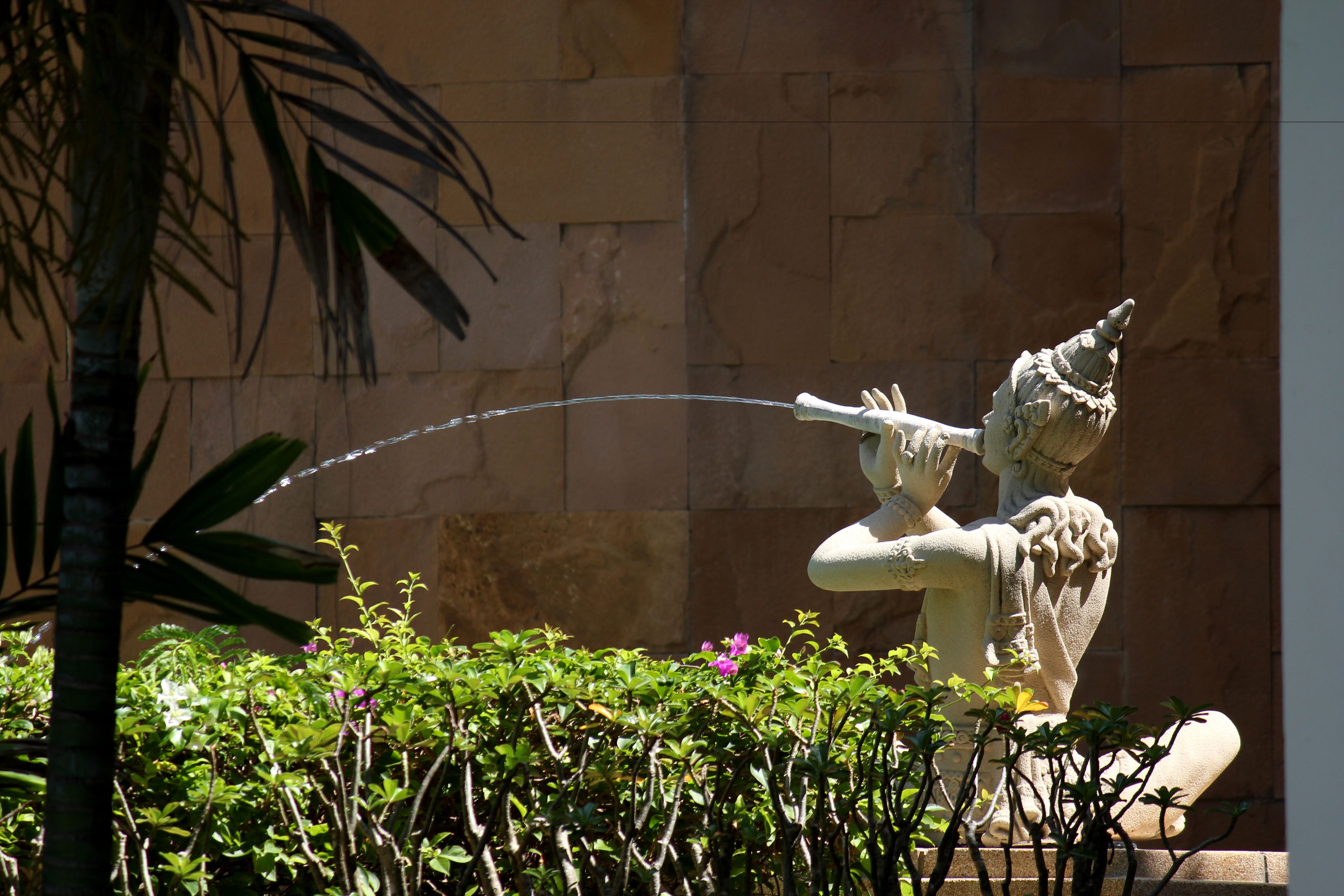 Una estatua de una figura con una lanza, frente a un muro de piedra y con vegetación al frente.