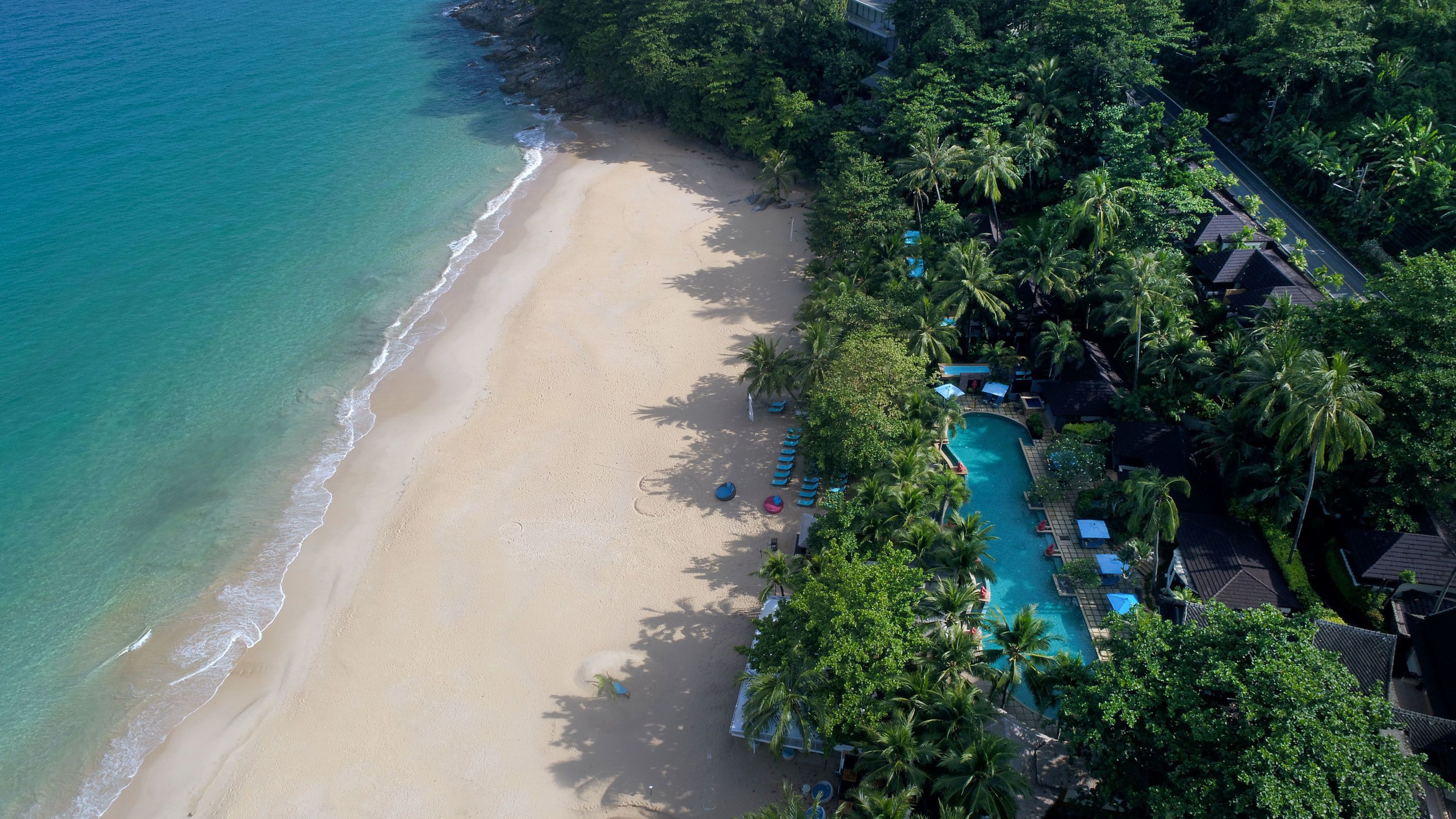 An aerial view of a beach resort with a swimming pool, beach, and lush greenery.
