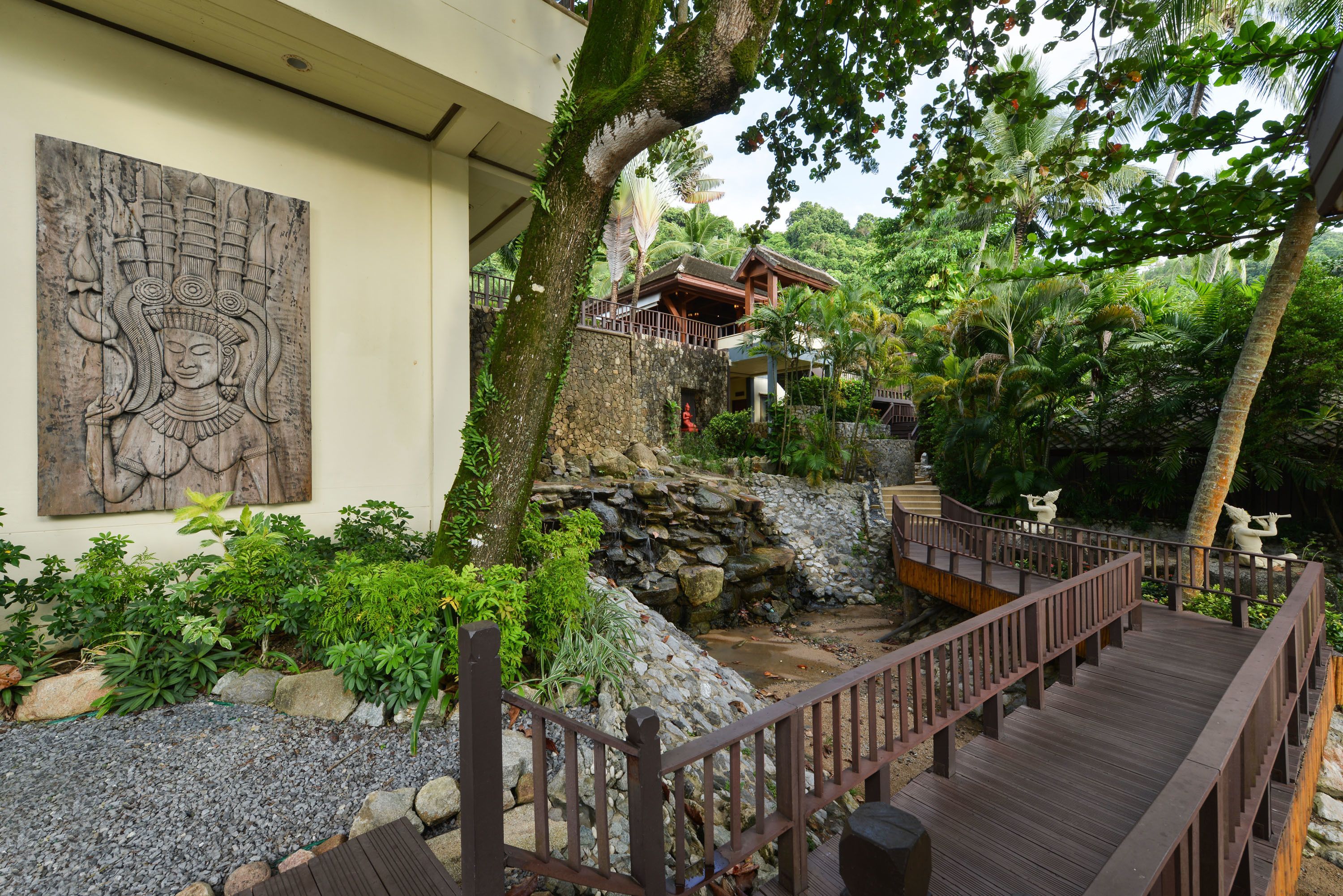 A wooden deck with a stone wall and a statue, surrounded by lush greenery and a traditional building with a mural.