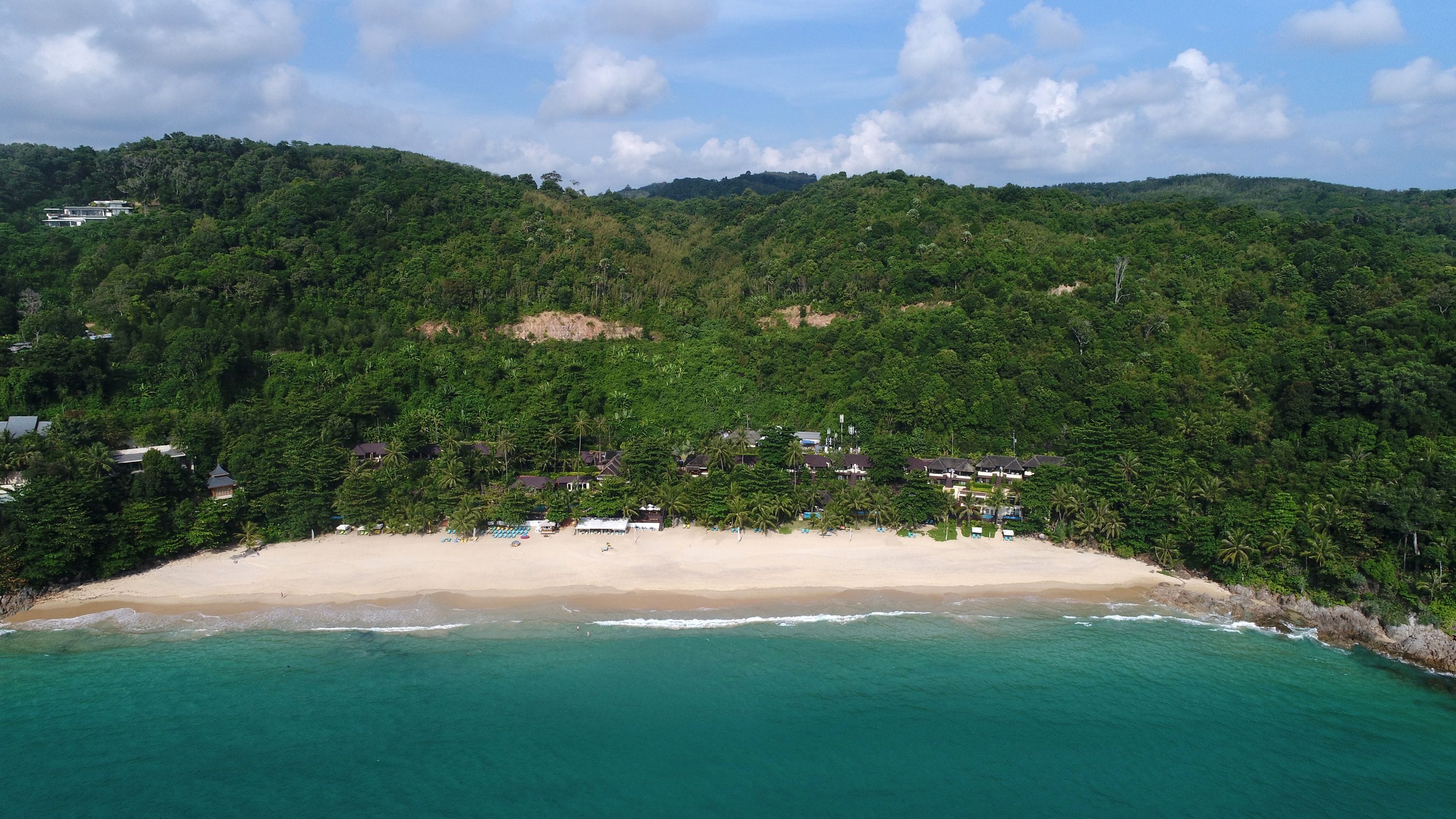 Una zona costera con una playa de arena, aguas turquesas cristalinas y colinas boscosas de color verde intenso al fondo.