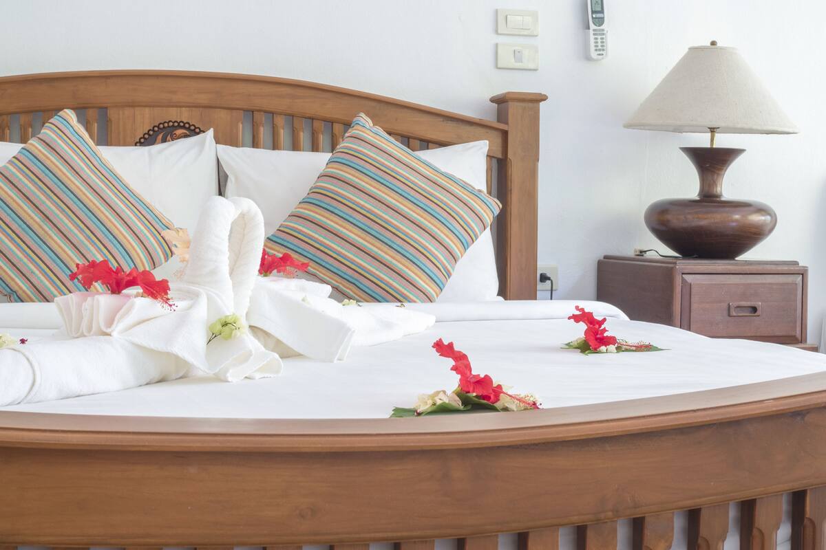 Cama bem arrumada com almofadas listradas, luminária de cabeceira e mesa de apoio com telefone e interruptor.