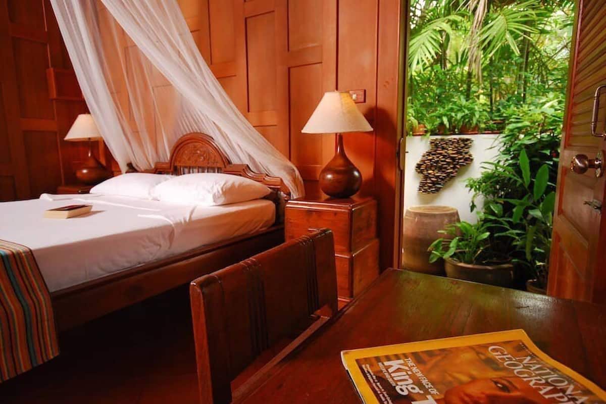 Quarto com cama, móveis de madeira, uma mesa com revista e vista para o verde lá fora.