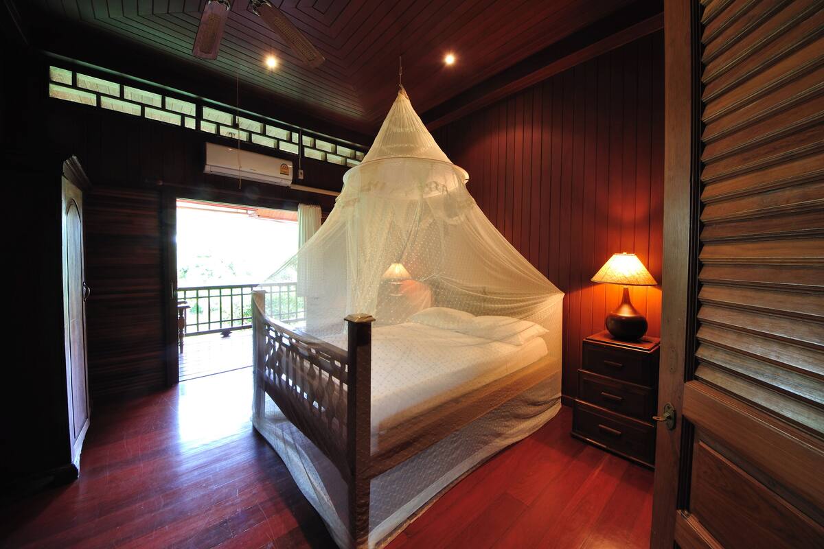 Quarto com cama de madeira, mesa de cabeceira com abajur e vista para o exterior pela janela.