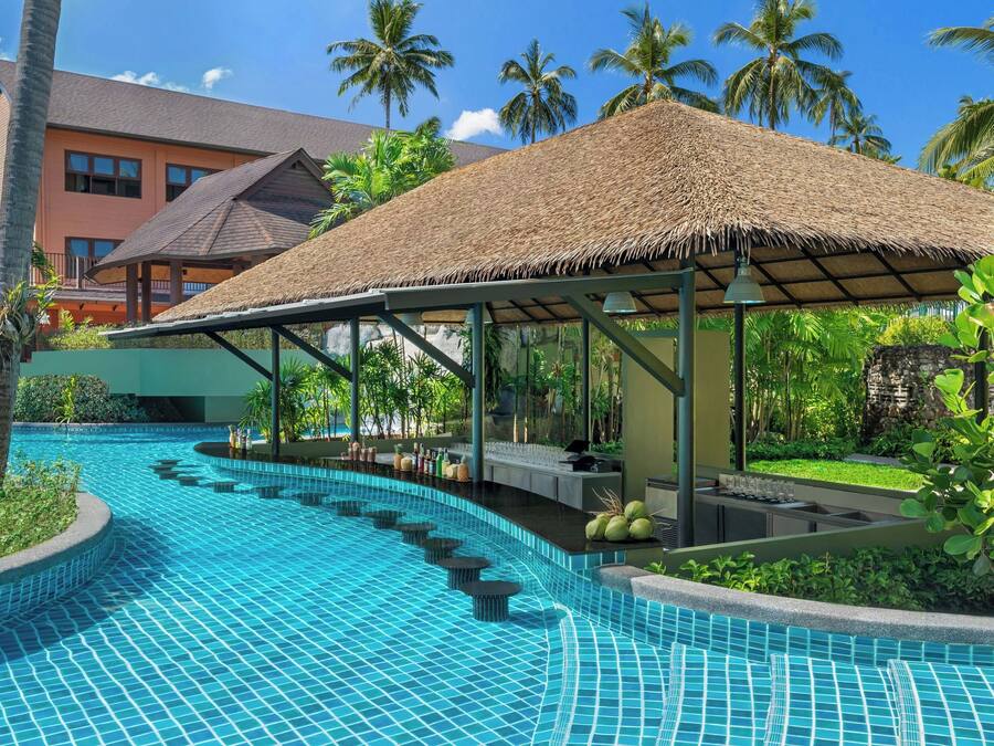 A swimming pool with a thatched roof seating area, surrounded by palm trees and a building with a red facade.