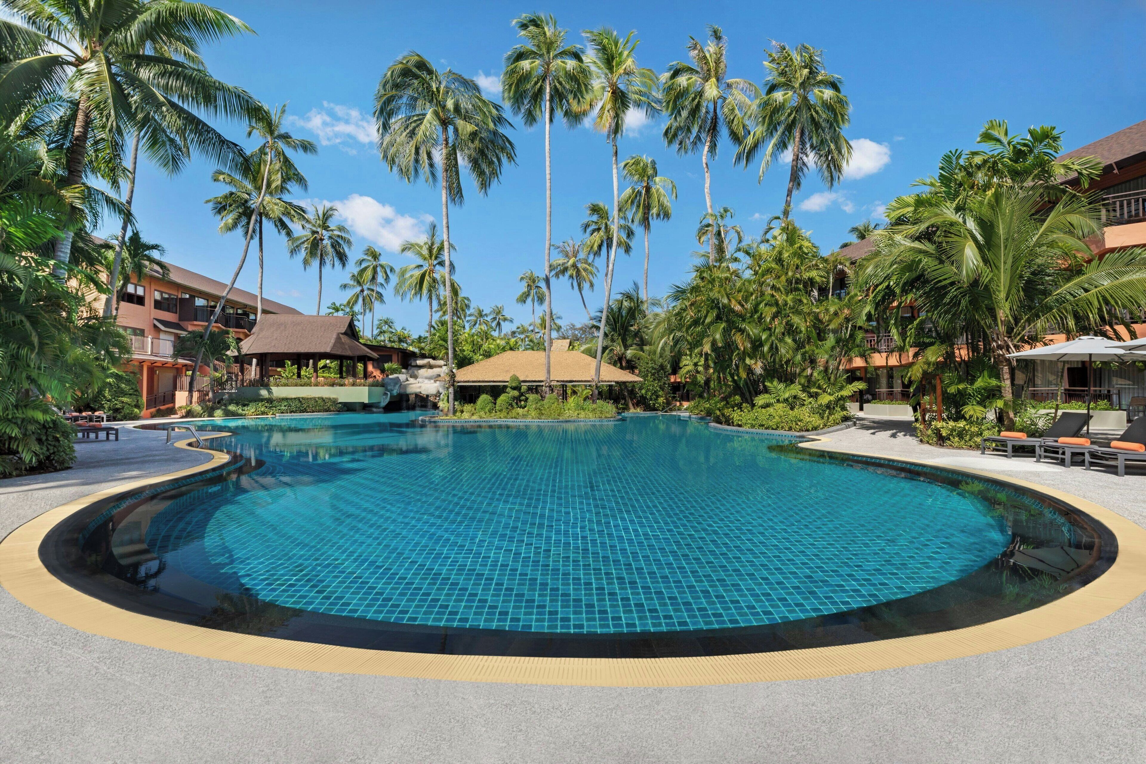 A large, clear swimming pool surrounded by palm trees and hotel buildings.
