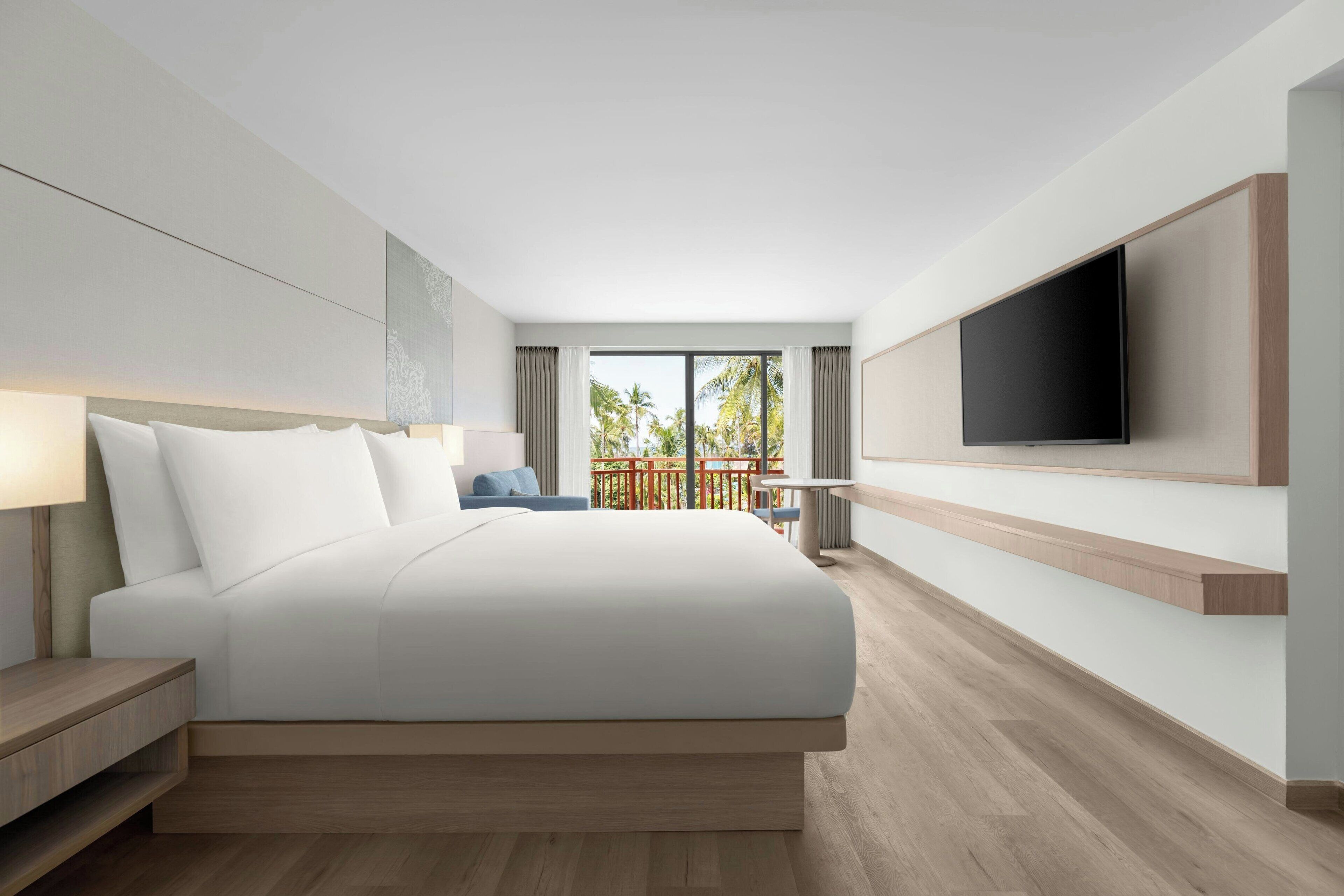 A modern hotel room with a large bed, a flat-screen TV mounted on the wall, a balcony with a view of palm trees, and a wooden floor.