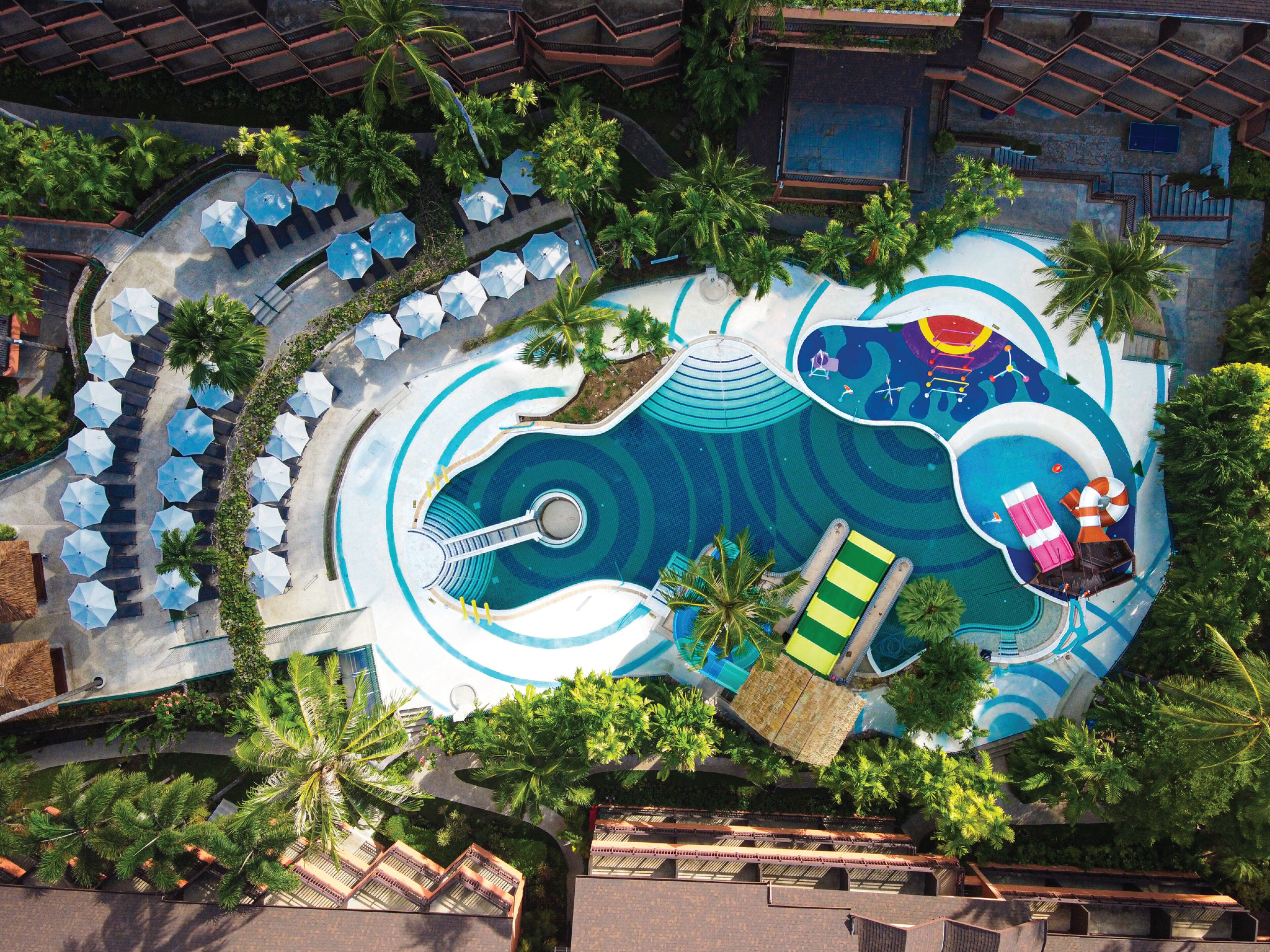 Vista aérea de un complejo turístico con un amplio área de piscina, numerosas tumbonas y palmeras.