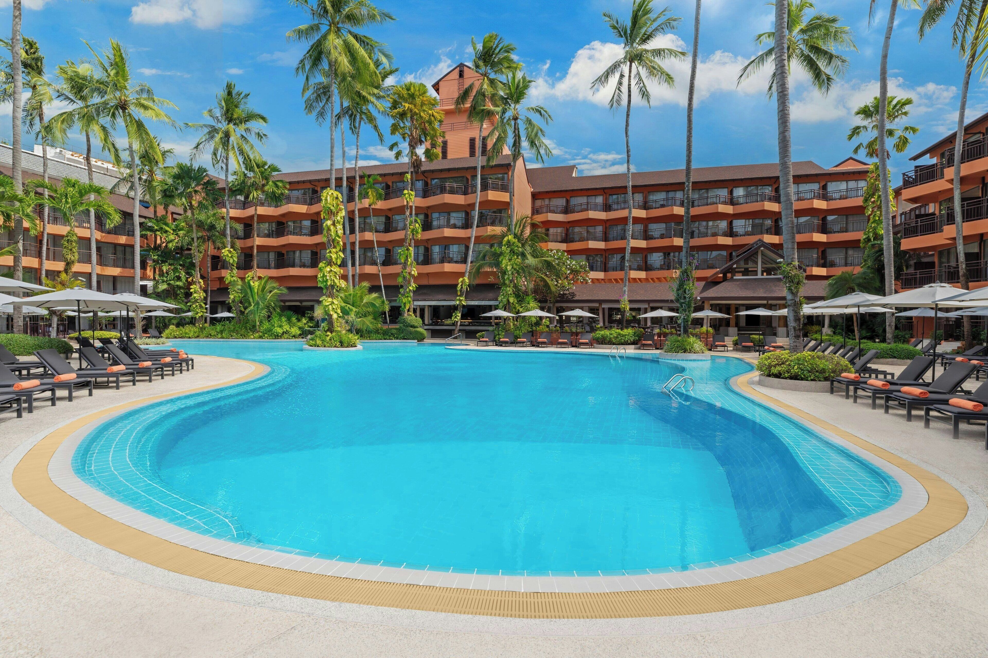 Una gran piscina rodeada de palmeras y sillas de descanso, con un edificio de hotel de varios pisos al fondo.