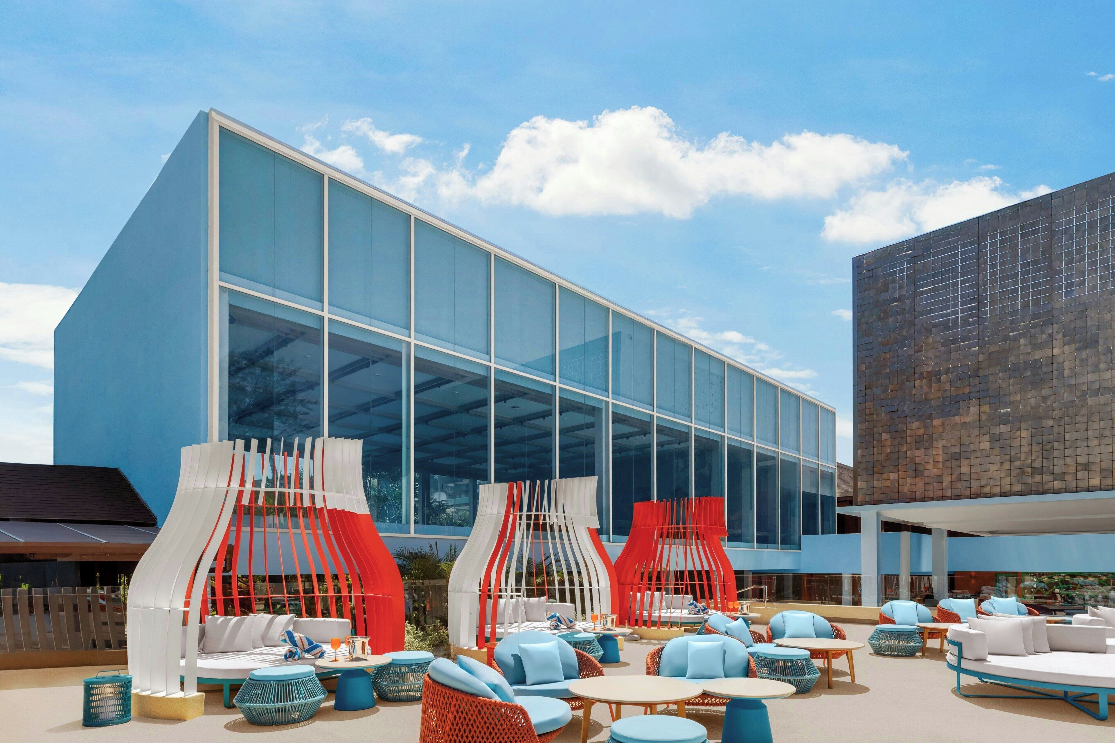 Un área de estar al aire libre moderna con estructuras blancas y rojas, mobiliario azul y blanco, y un gran edificio de cristal al fondo.