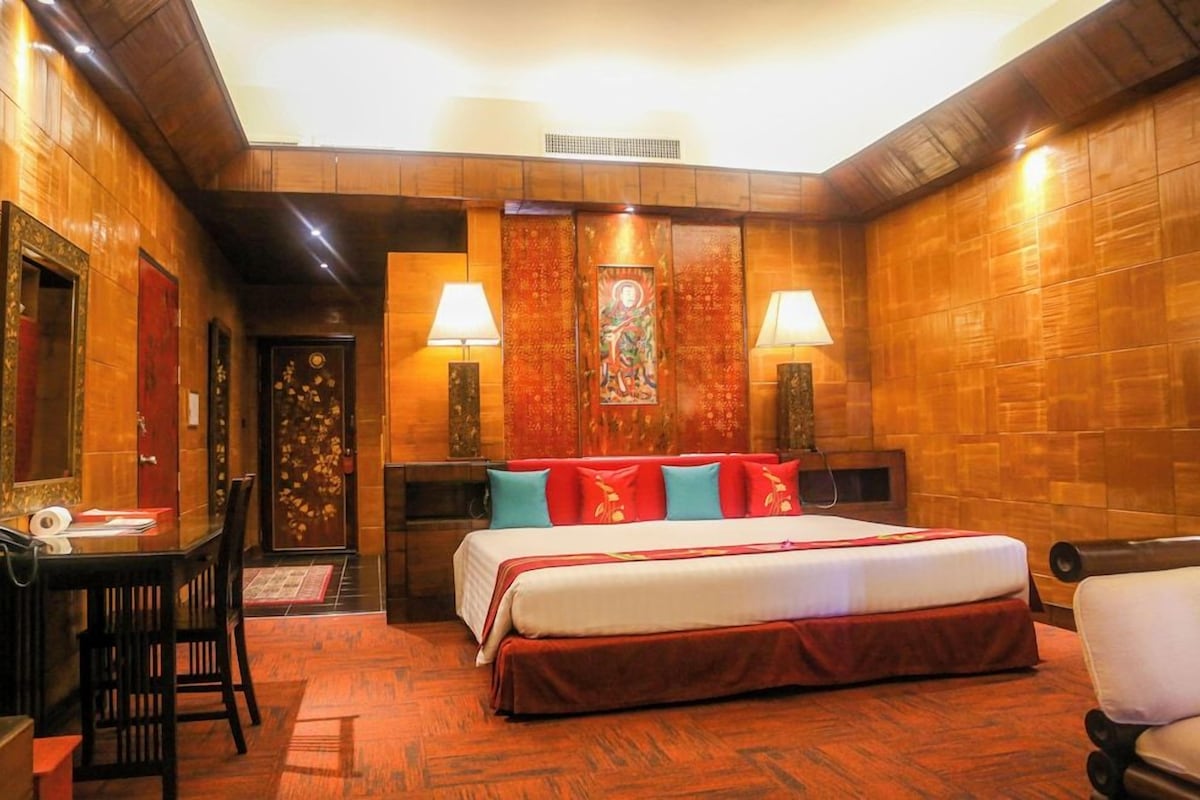 A hotel room with a wooden interior, a bed with red and white bedding, two bedside lamps, a desk with a chair, and a door with a floral design.