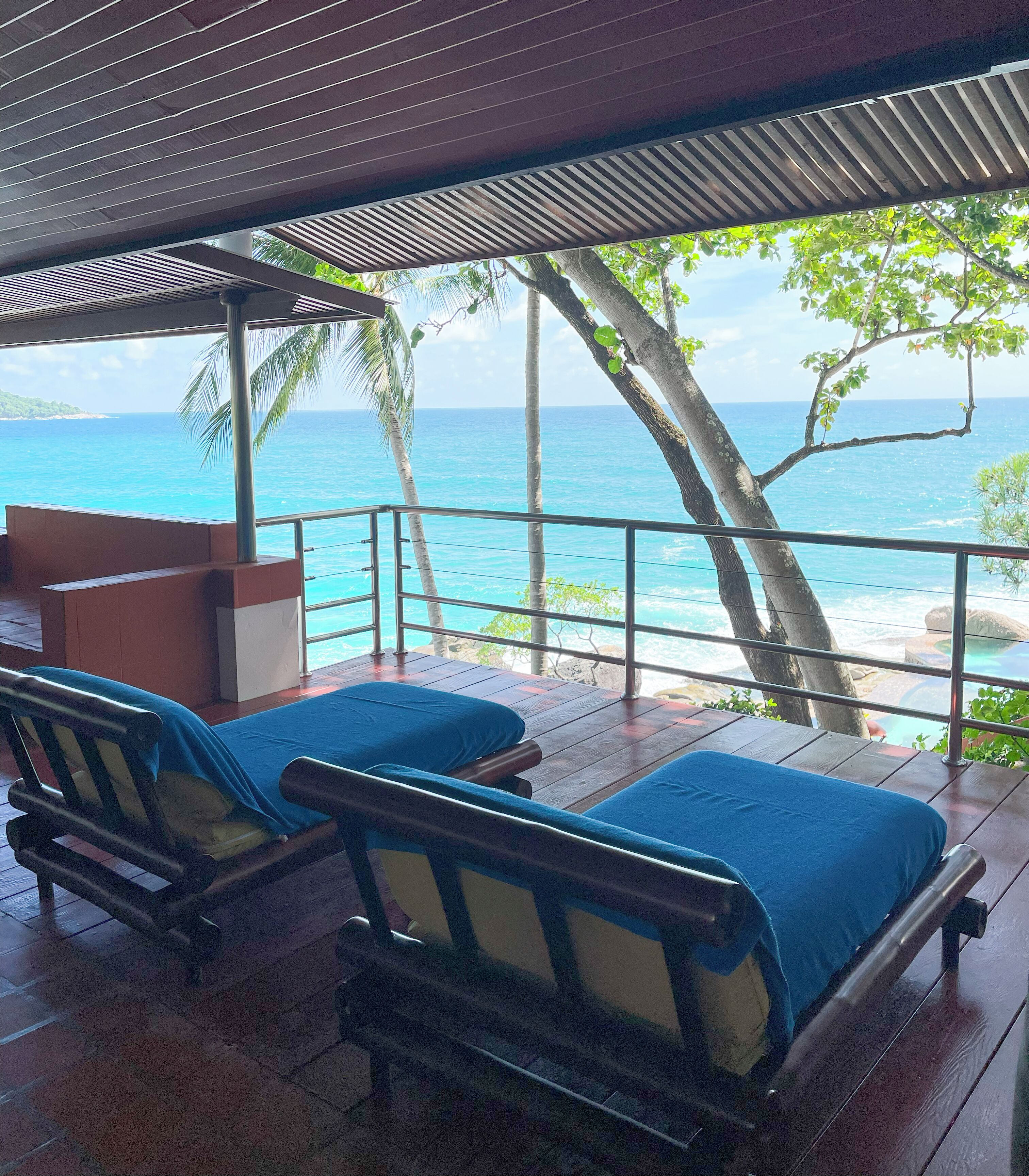 A covered outdoor seating area with two wooden benches, each with a blue cushion. The area overlooks a beach with clear blue water and palm trees.