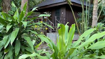 A wooden house with a thatched roof, surrounded by lush greenery and a sign indicating 'BAAN KRATING 127-129'.