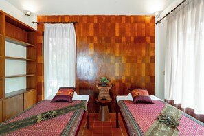 A room with two beds, wooden paneling on the walls, a small table with a plant, and a shelf unit.