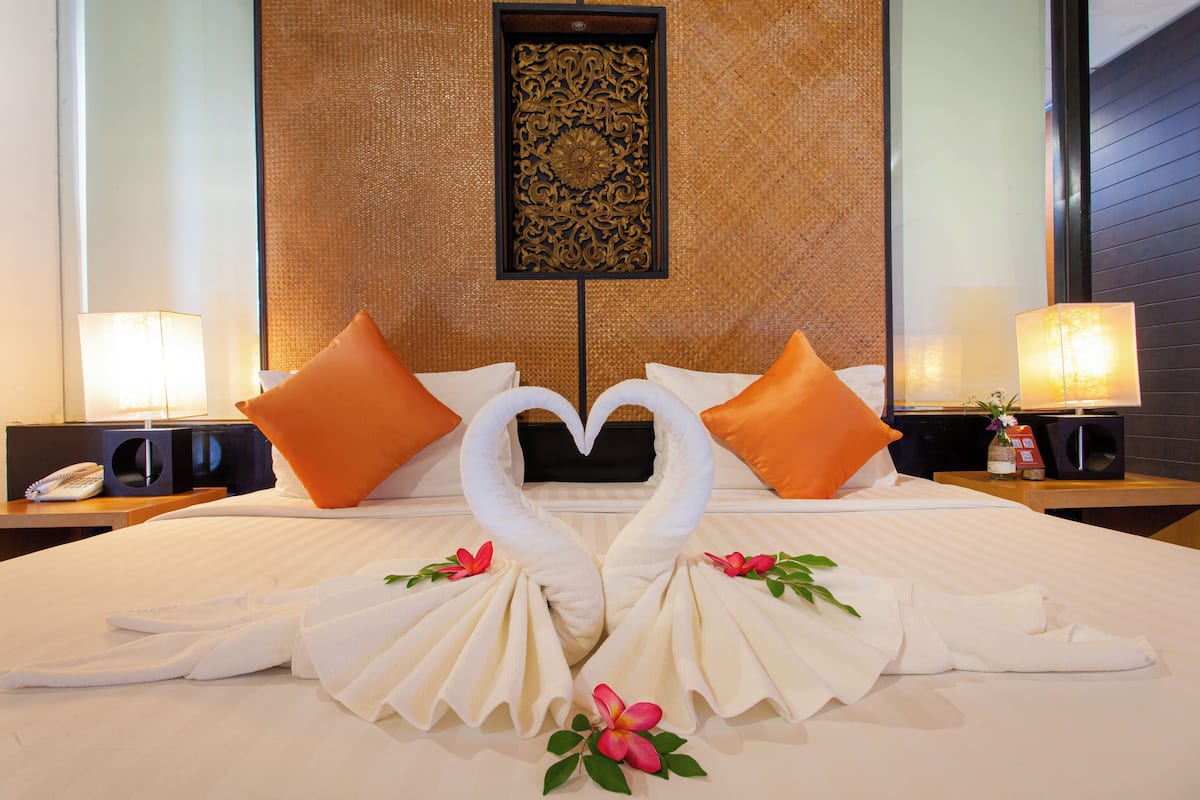 Habitación de hotel con una cama decorada con una toalla dispuesta en forma de corazón y almohadas naranjas.