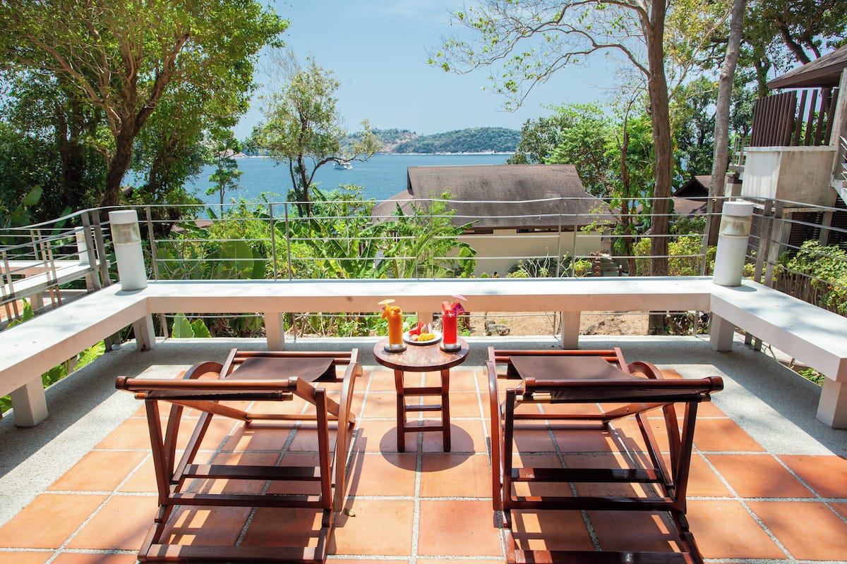 Una terraza con dos sillas de madera y una mesa, con vista a un cuerpo de agua y follaje.