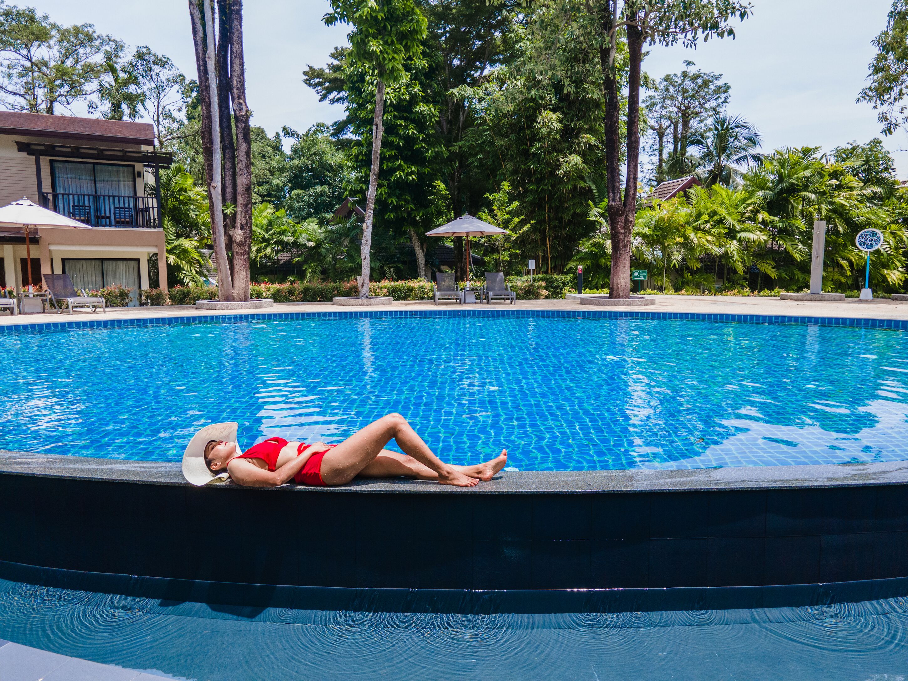 A pool with a person lying on the edge, surrounded by trees and a building in the background.