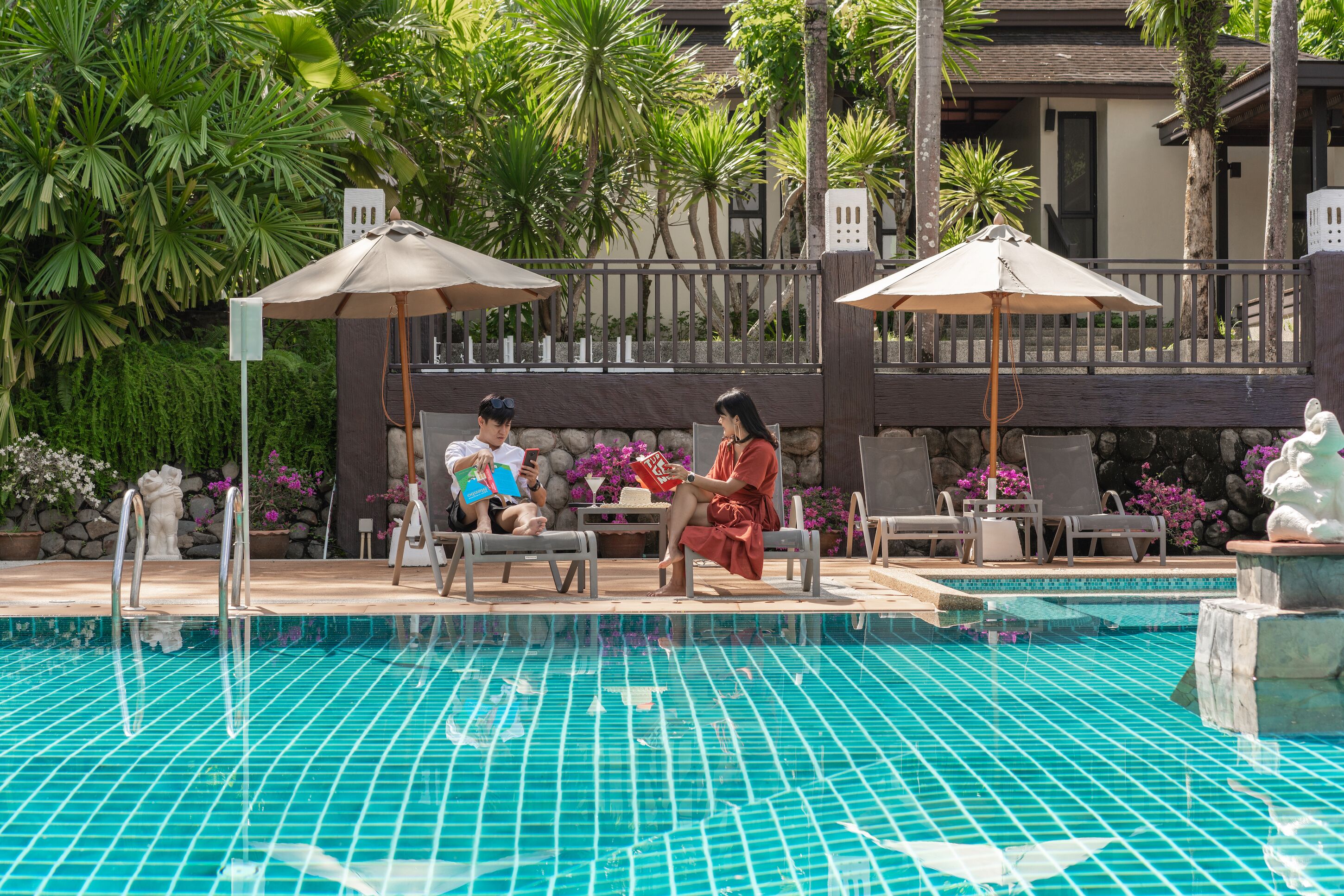 Un espace aménagé au bord de la piscine, avec des chaises longues et des parasols, entouré d’une végétation luxuriante et d’un mur en pierre.