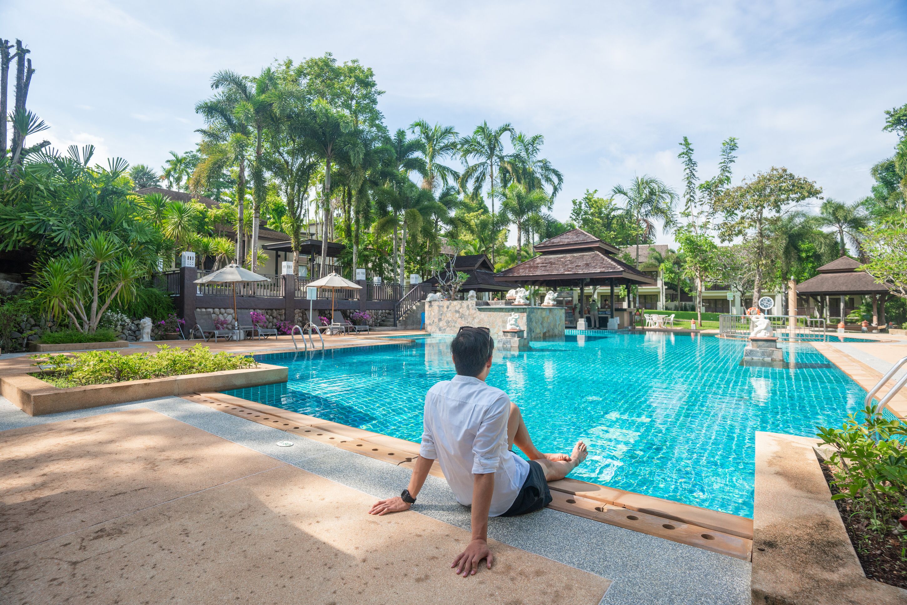 Un homme est assis au bord d’une piscine, avec un jardin tropical et des espaces de détente en arrière-plan.