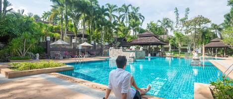 Un homme est assis au bord d’une piscine, avec un jardin tropical et des espaces de détente en arrière-plan.