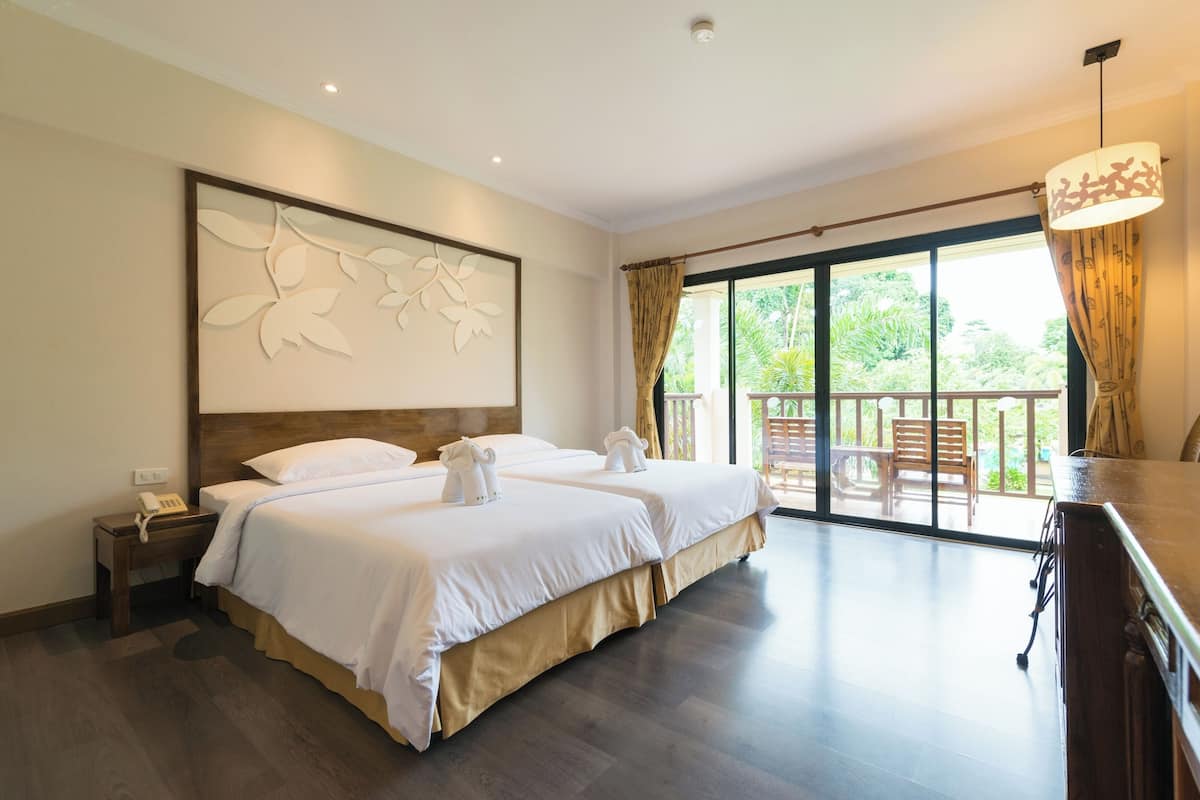 Habitación de hotel con dos camas, cabecero de madera, un ventanal grande con cortinas, suelo de madera y una mesita con un teléfono.