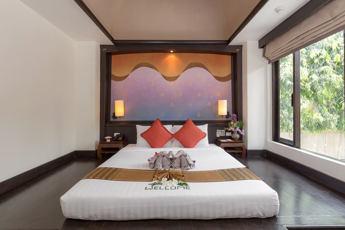 A hotel room with a large bed, a welcome mat, and a view of greenery through the window.