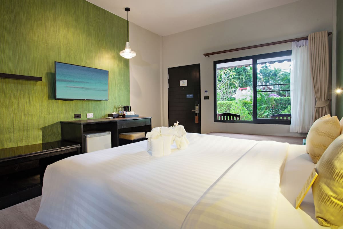 A hotel room with a large bed, a desk, a television, and a window with a view of greenery.
