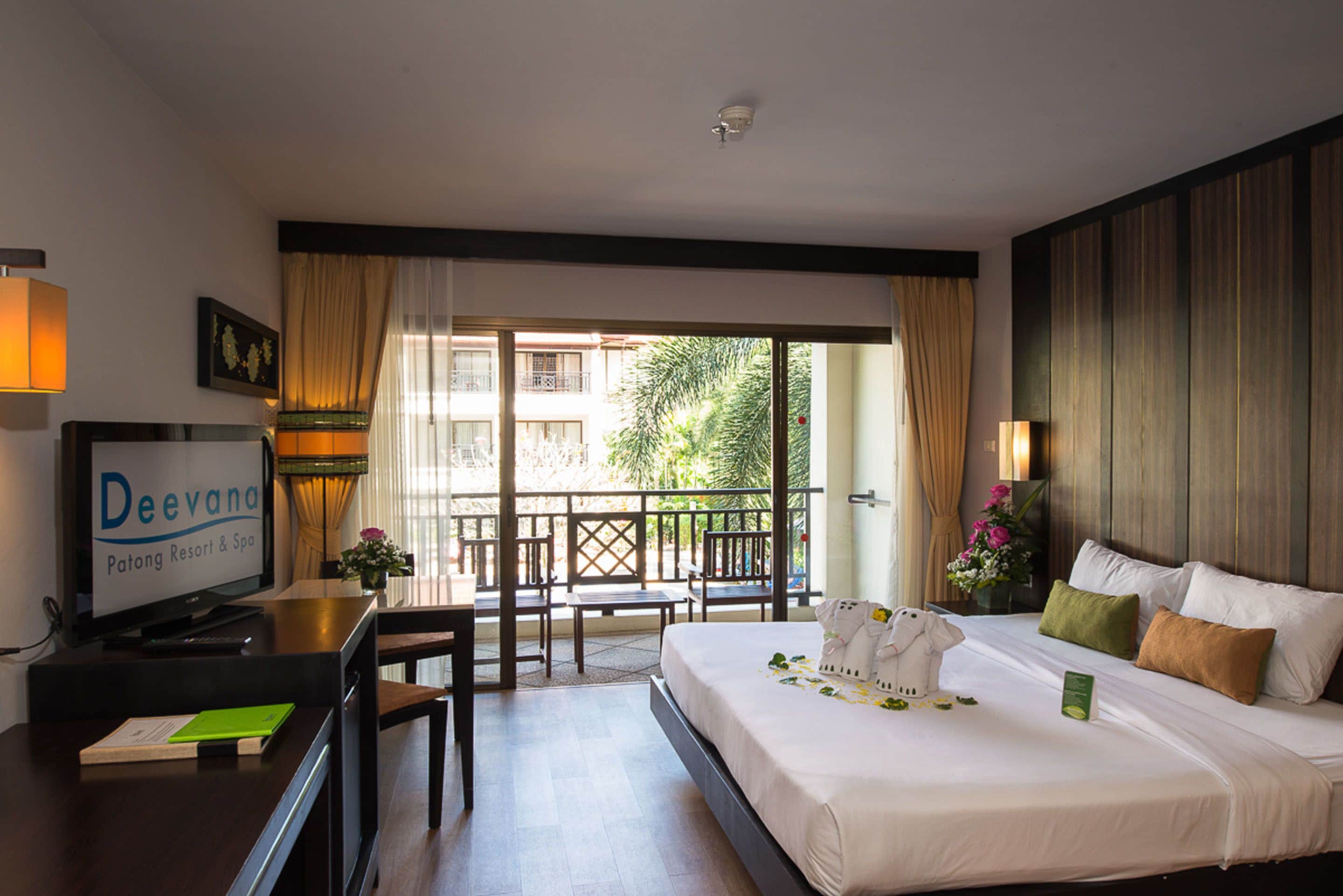 A hotel room with a bed, a desk with a television, a balcony with chairs, and a view of greenery outside.