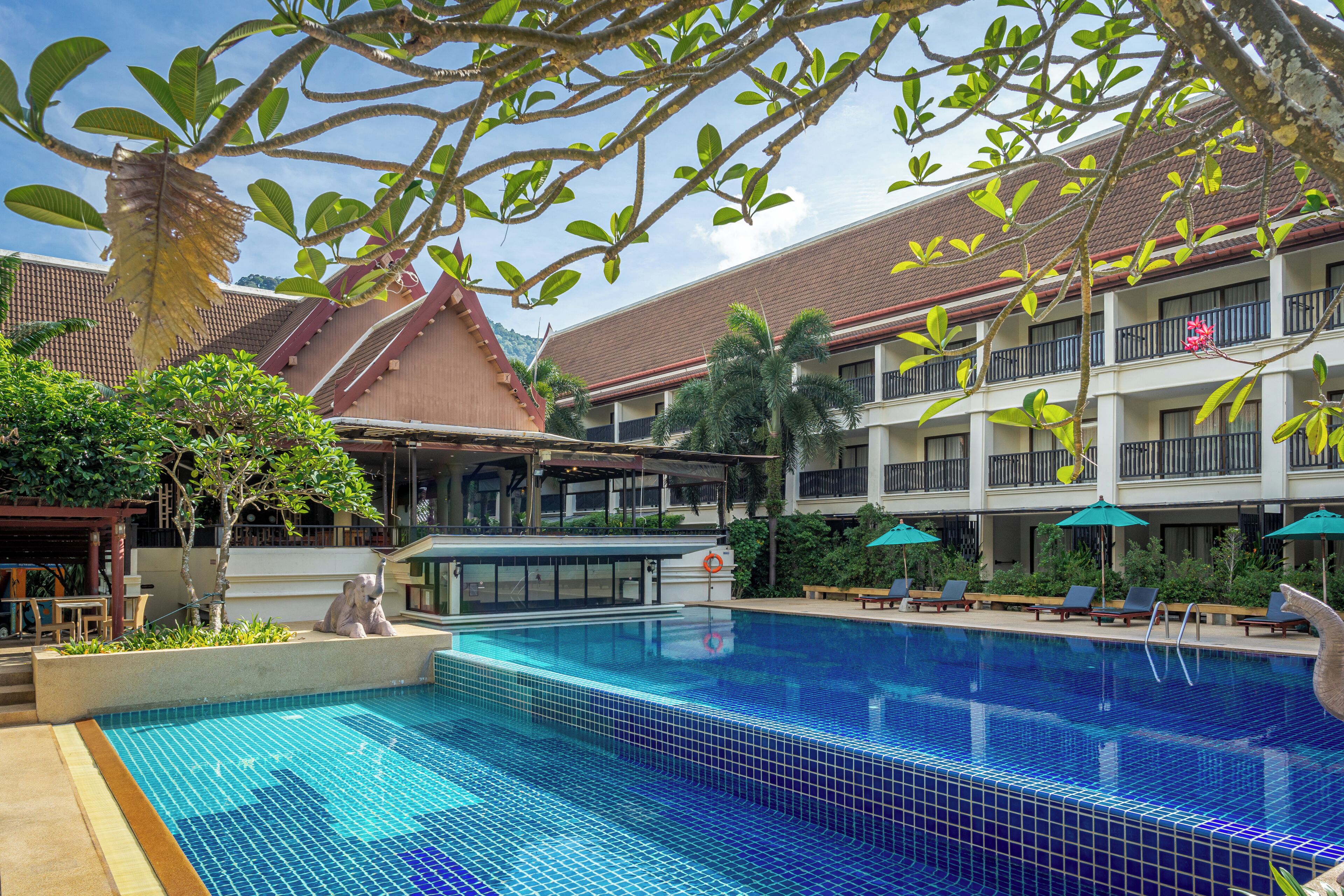 Piscina com deck de azulejos, rodeada por espreguiçadeiras e guarda-sóis, com um edifício com varandas ao fundo.