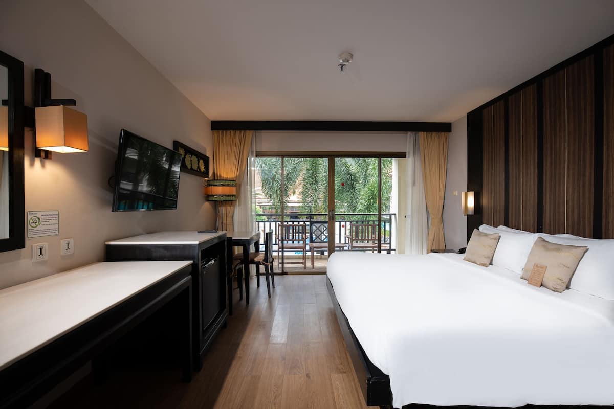 A hotel room with a bed, a desk, a chair, a TV, and a balcony with a view of trees.