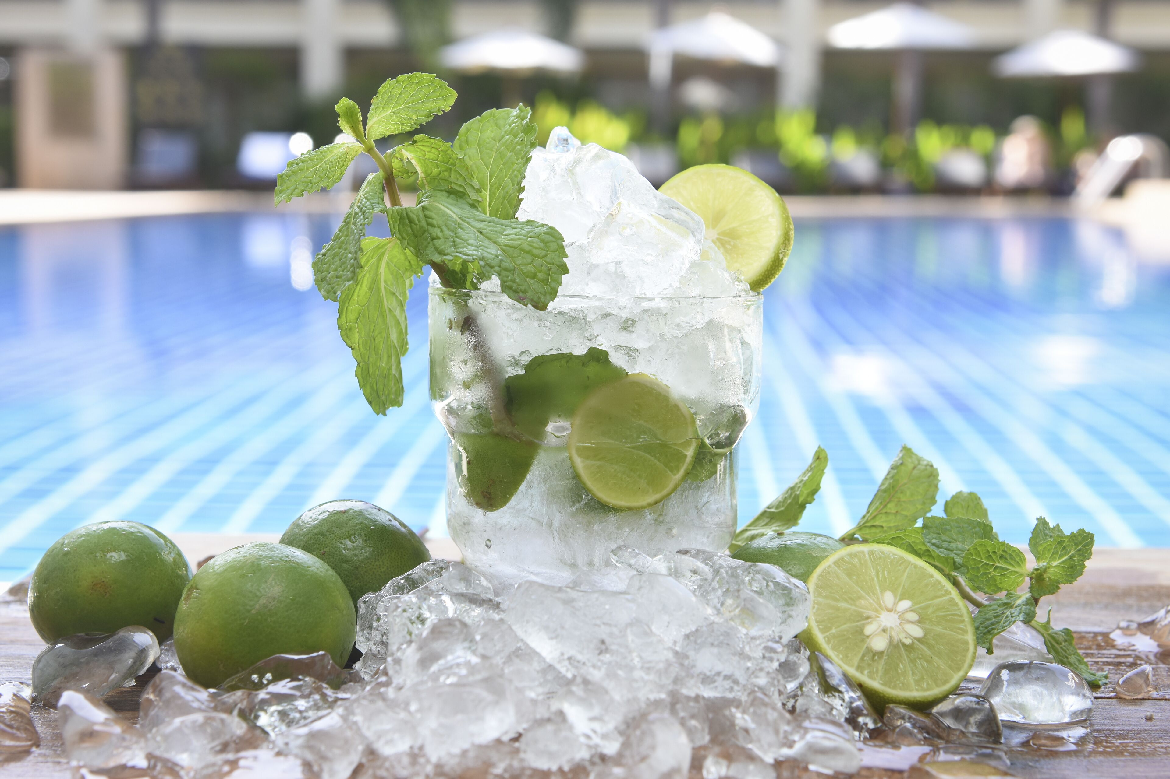 A glass of a refreshing drink with ice and lime, surrounded by additional limes and mint leaves, placed on a poolside setting.